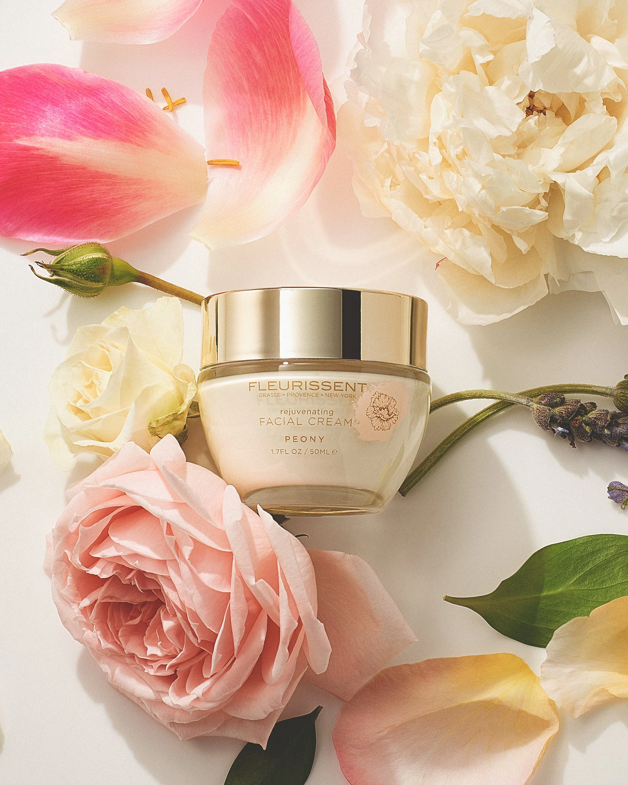 Peonies roses and lavender frame up a jar of luxury face cream on a white surface with bright, open shadows