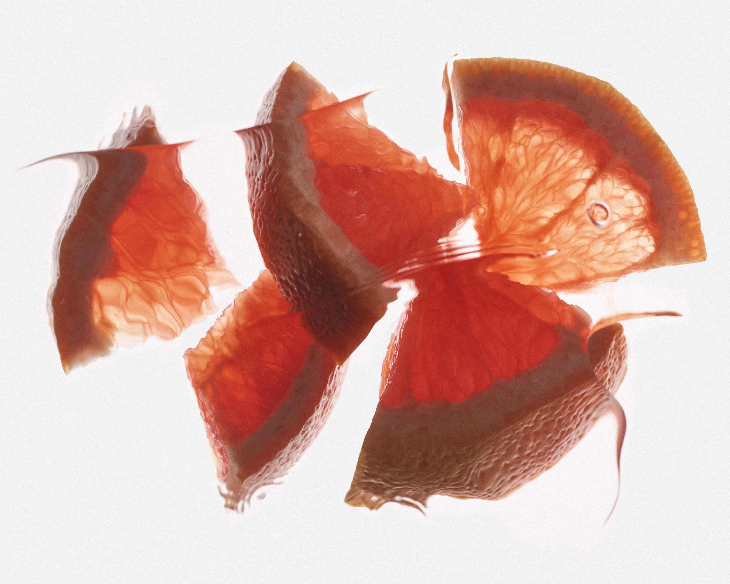 Close-up of thinly sliced red and orange grapefruit pieces, against a white background with water ripples and bubbles above the fruit pieces