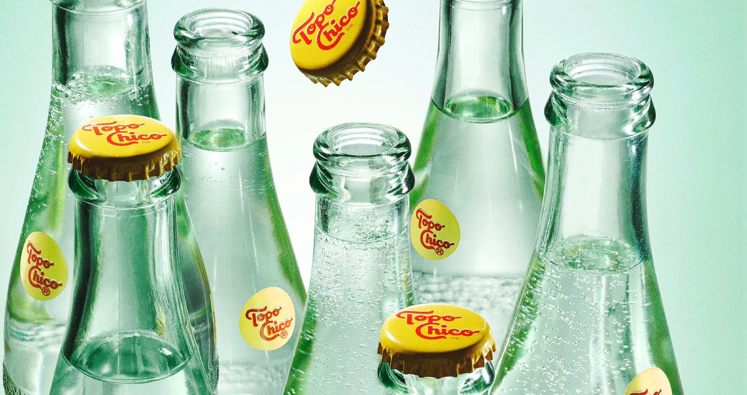 Multiple glass soda bottles with yellow caps labeled 'Topo Chico' on a light green background.