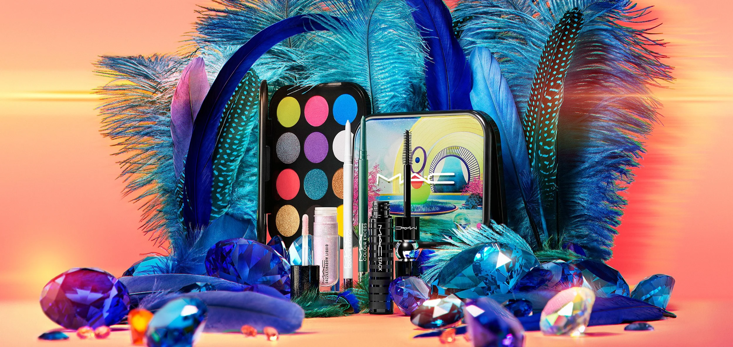 Colorful makeup products including eyeshadow palette, lip gloss, mascara, and various brushes arranged with large blue feathers and numerous purple and blue gemstones, set against a warm gradient background.