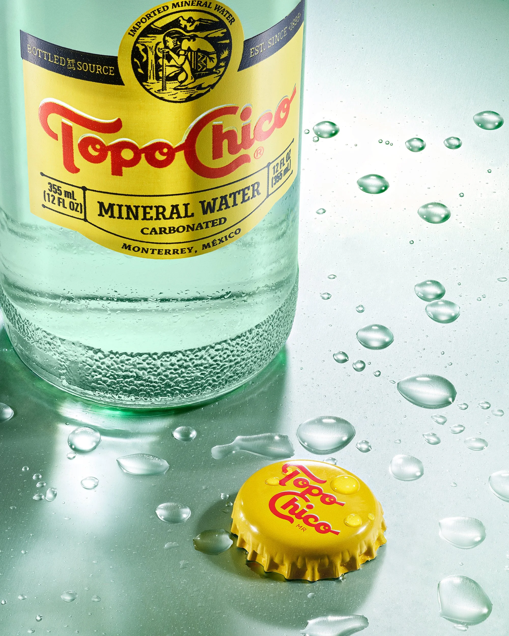 A single glass bottle of mineral water withthe name Topo Chico stands next to its metal bottle cap on a reflective surface that is covered in water droplets