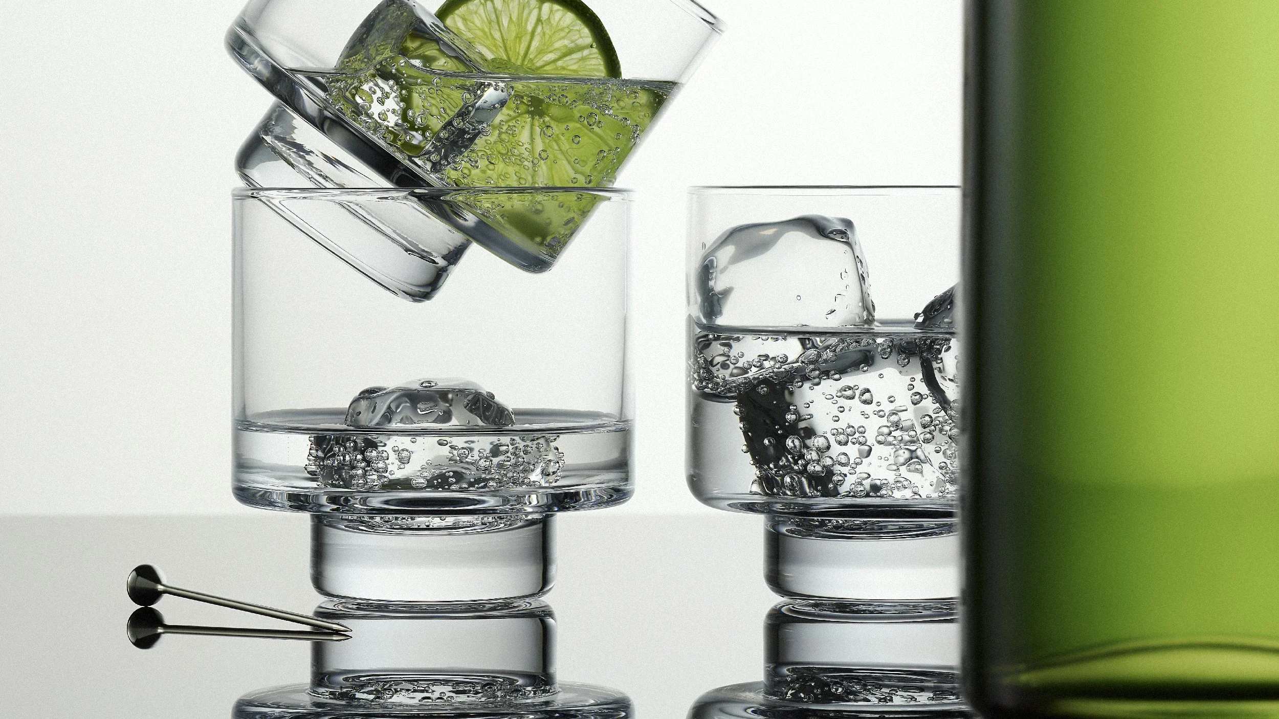 Three glasses of sparkling water with ice cubes, garnished with lime slices. One glass is tilted, pouring water into the middle glass. The glasses are on a reflective surface with a white background and a green object partially visible on the right side.