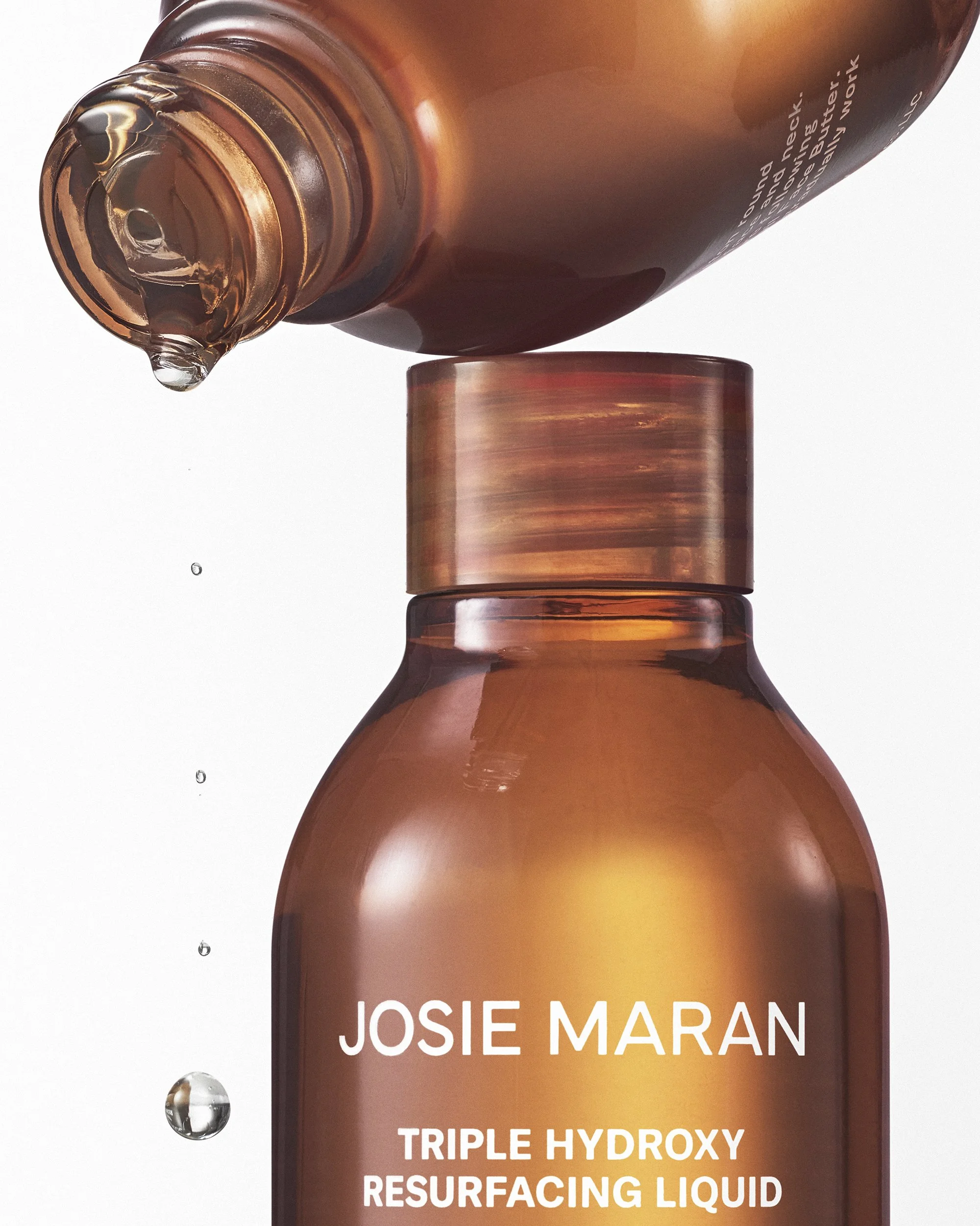 Josie Maran Triple Hyroxy amber colored skincare product drips from an open bottle while balancing on another bottle against a white background