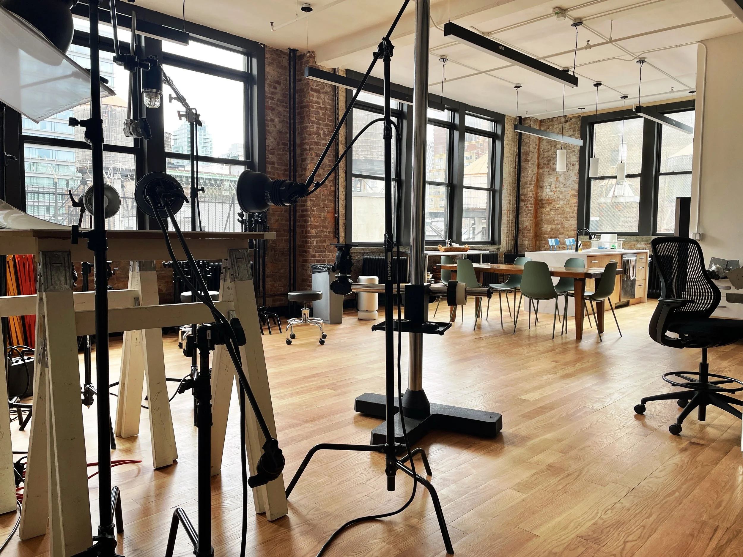 Empty modern coworking space with wooden floors, large windows, and a variety of chairs and tables, with some photography equipment visible in the foreground.