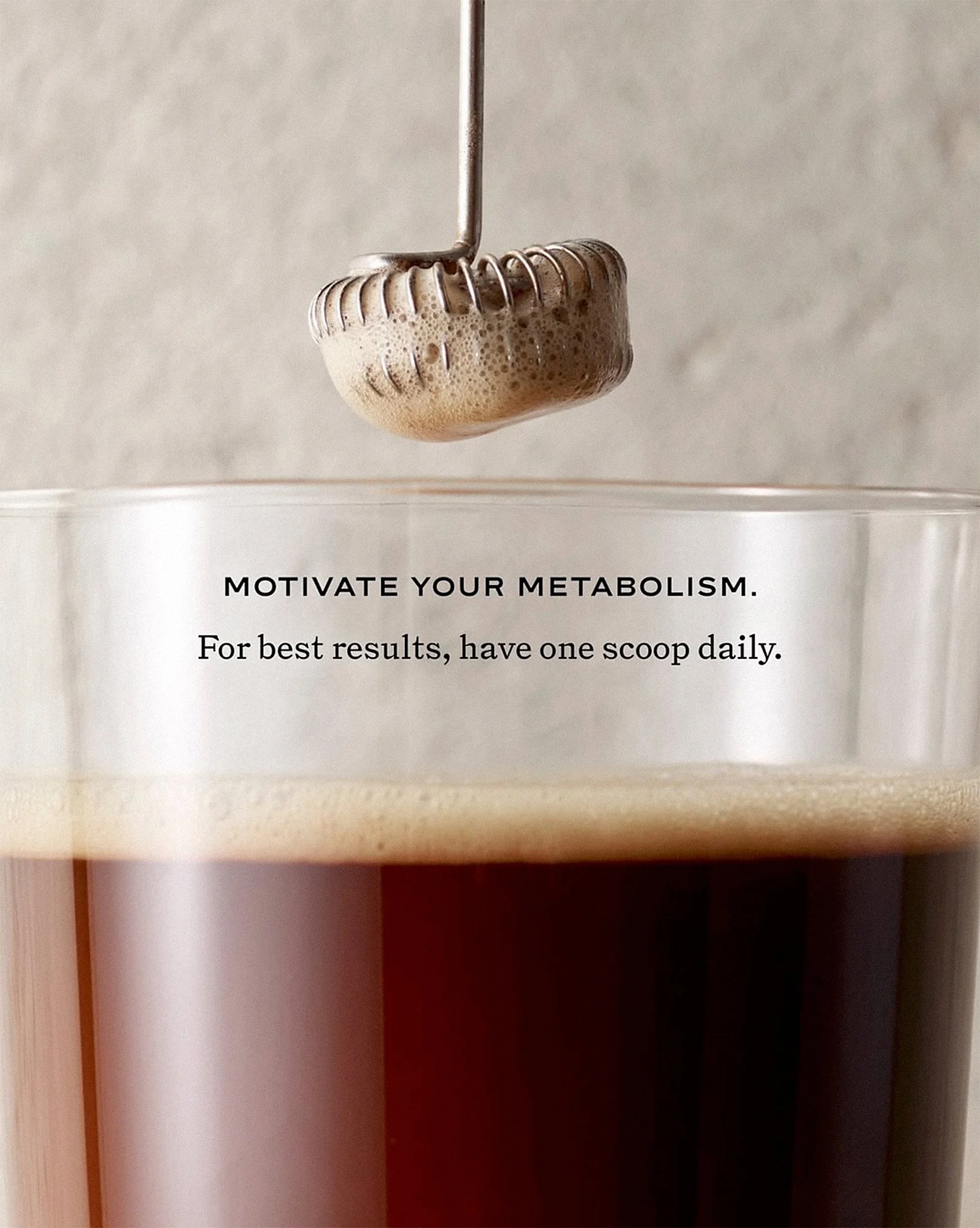 A closeup of a metal frother hovering over a drinking glass with a rich brown foamy beverage with a textured stone background