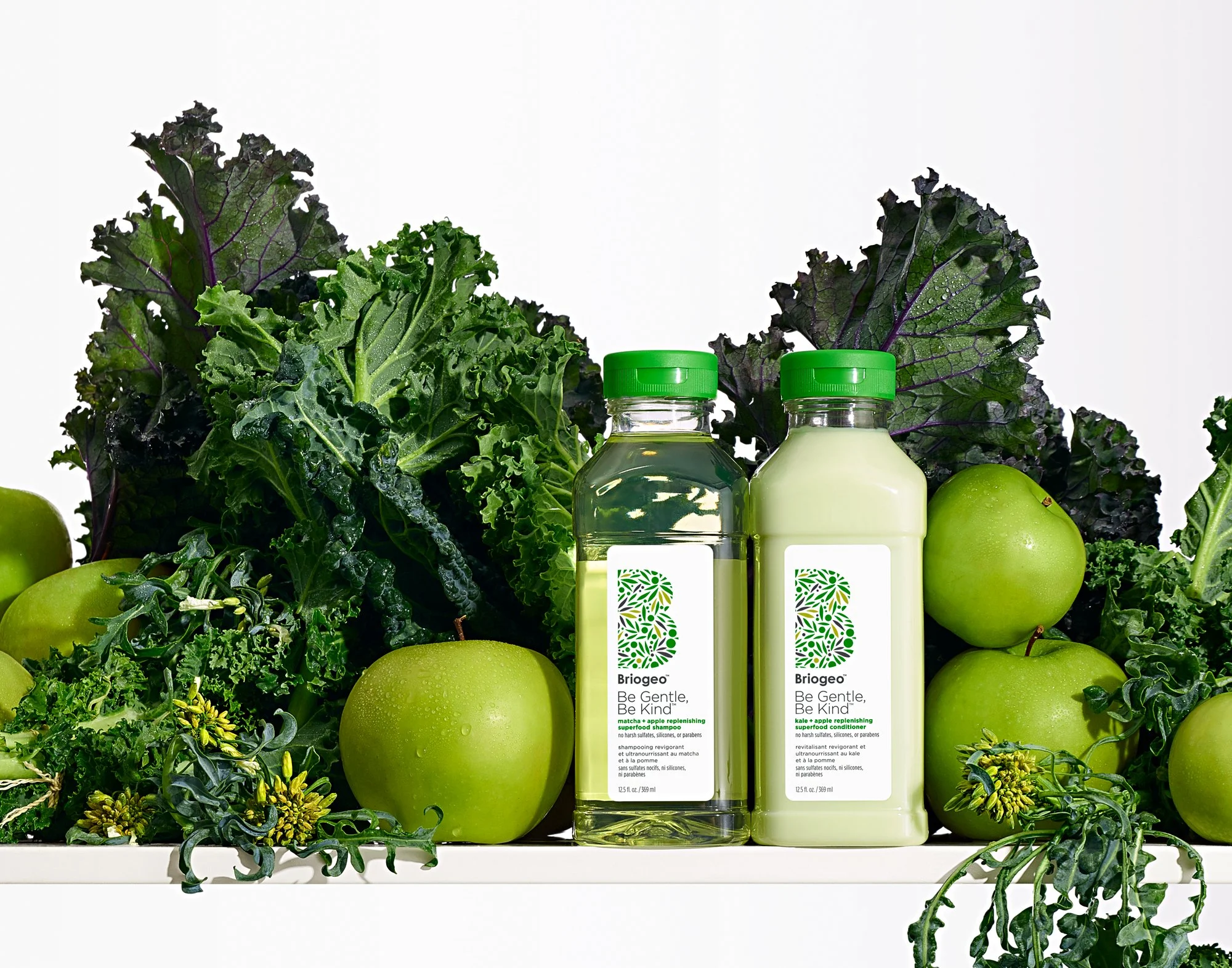 A shelf piled high with fresh kale and green apples surrounds two bottles of haircare product from the brand Briogeo