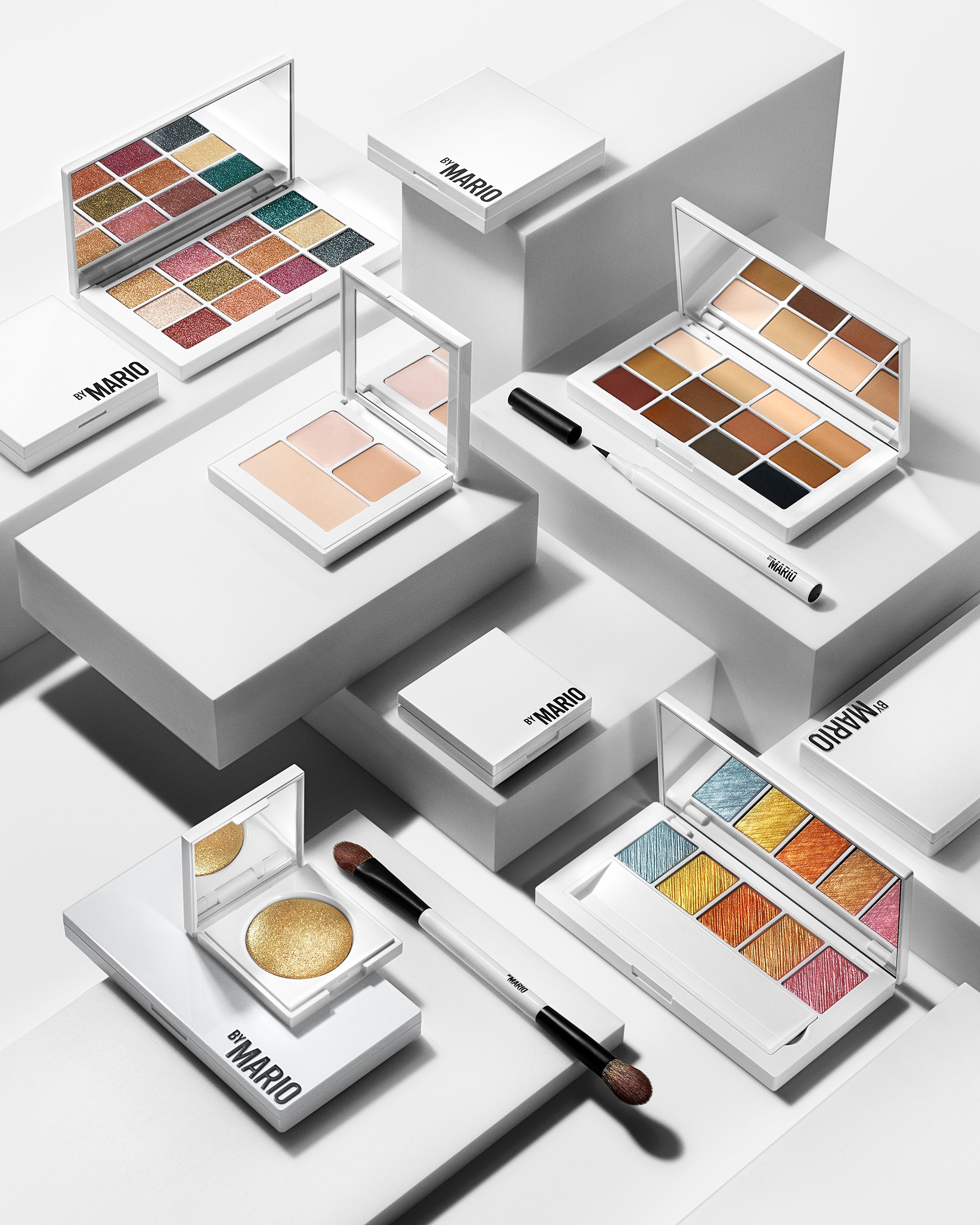 Makeup eyeshadow palettes, blush, and brushes on white platforms with shadows.