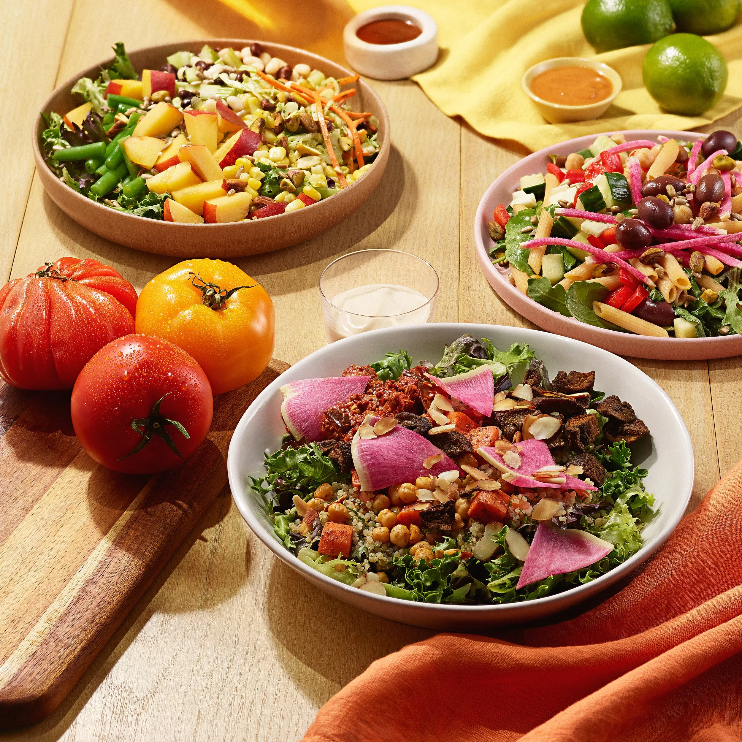 Three prepared plates of fresh salad appear on a wood table along with a trio of tomatoes, a glass with smoothie and orange linen napkin