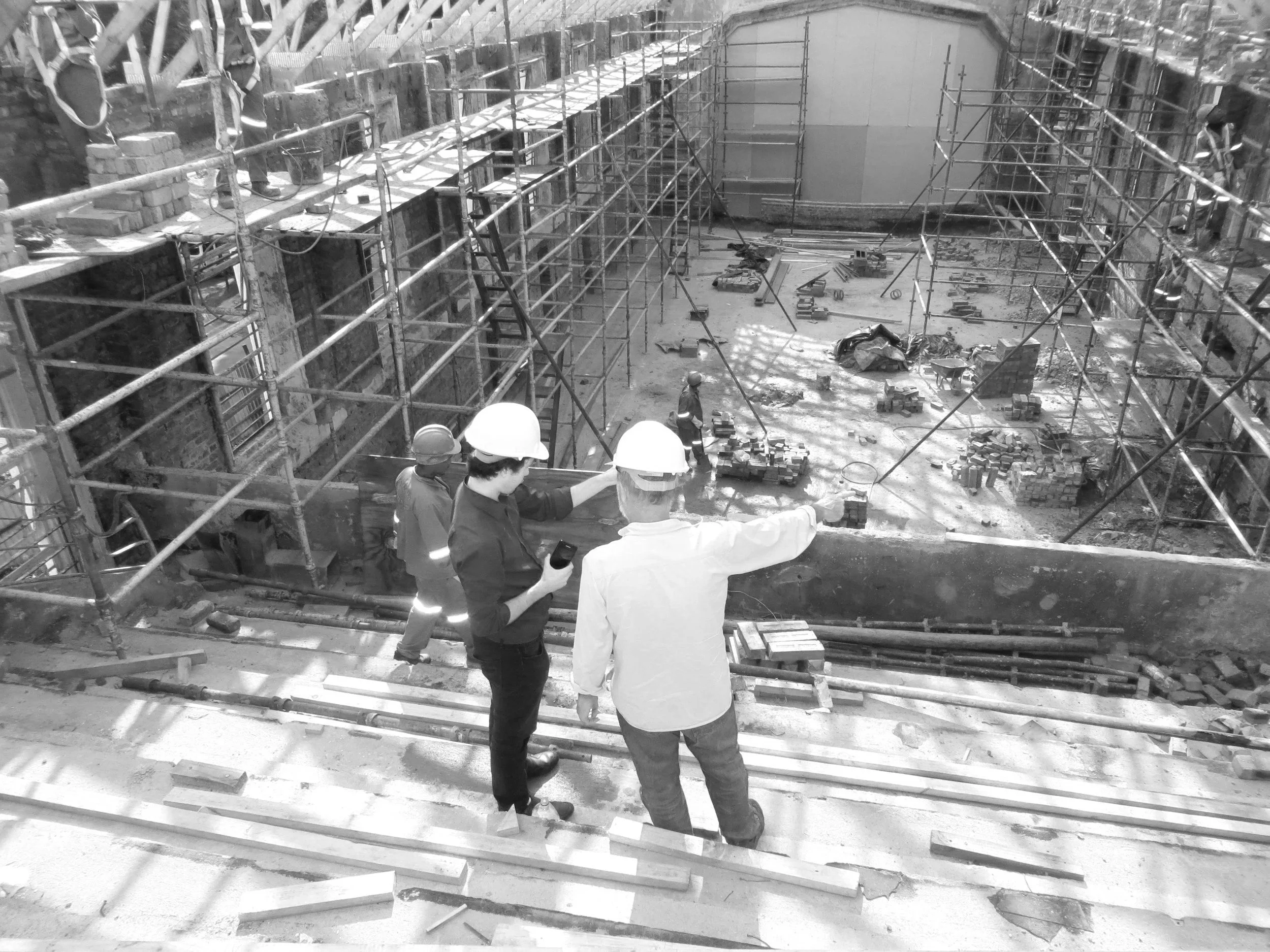 Construction site with scaffolding and workers, two people in the foreground wearing hard hats discussing, and other workers working on the site.