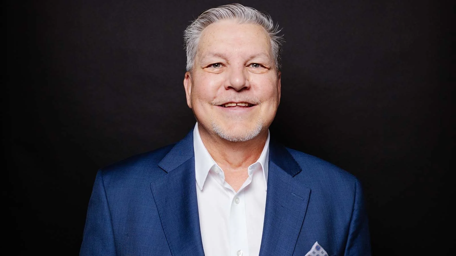 Portrait of a smiling older man in a blue suit and white shirt against a black background.