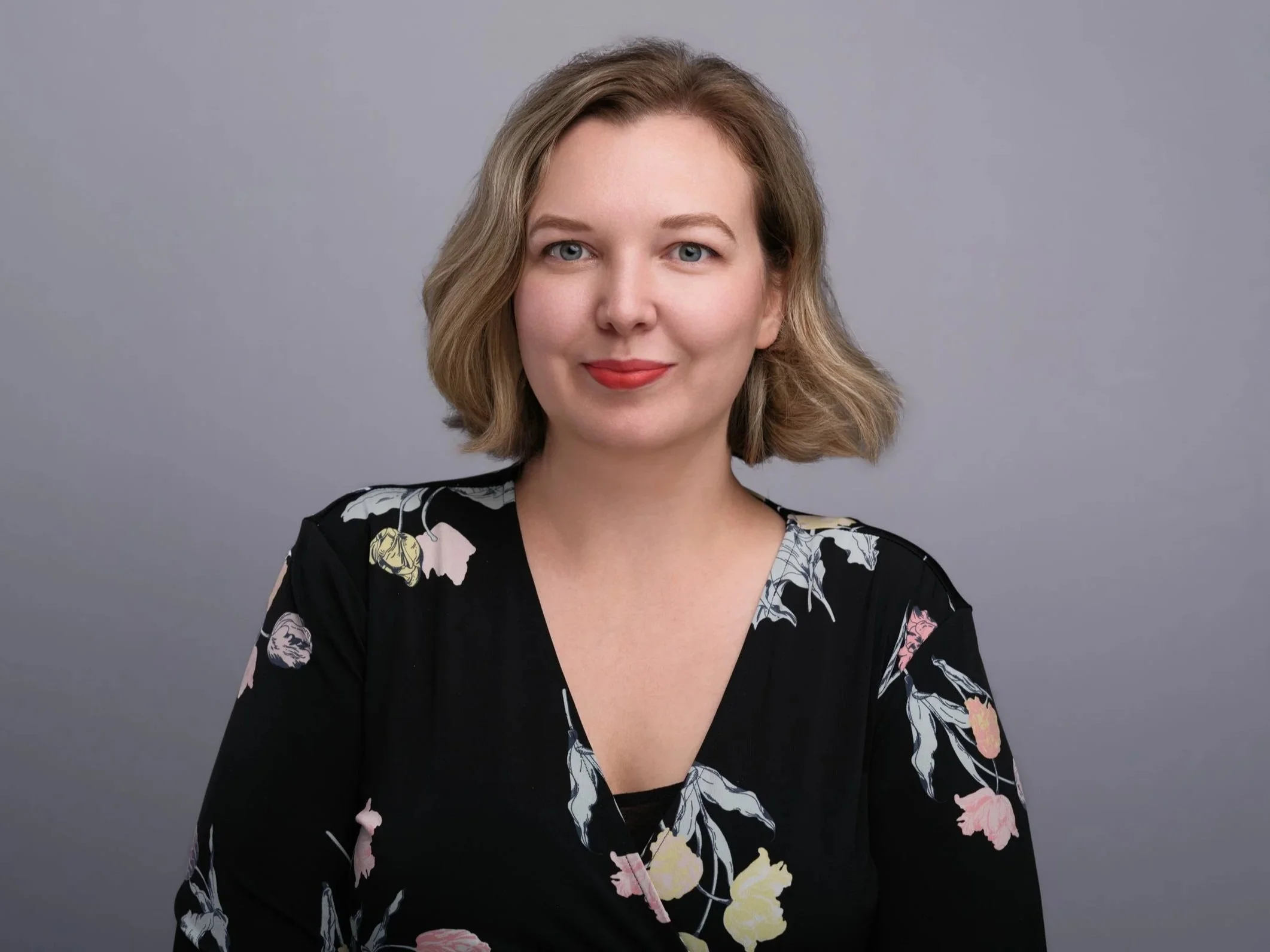 A woman with short, wavy blonde hair wearing a black dress with a floral pattern, smiling against a gray background.