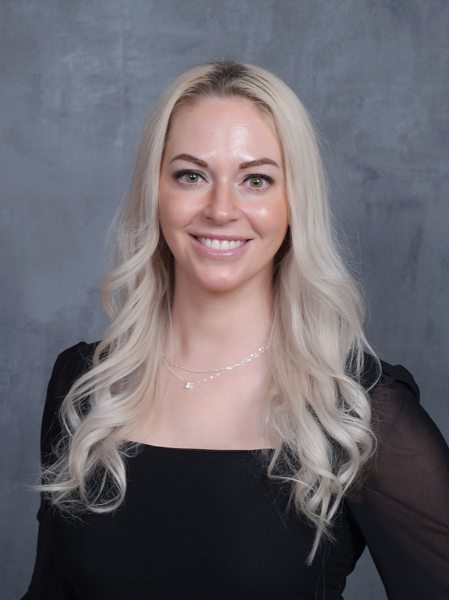 A woman with long blonde hair, wearing a black top, smiling, standing against a gray background.