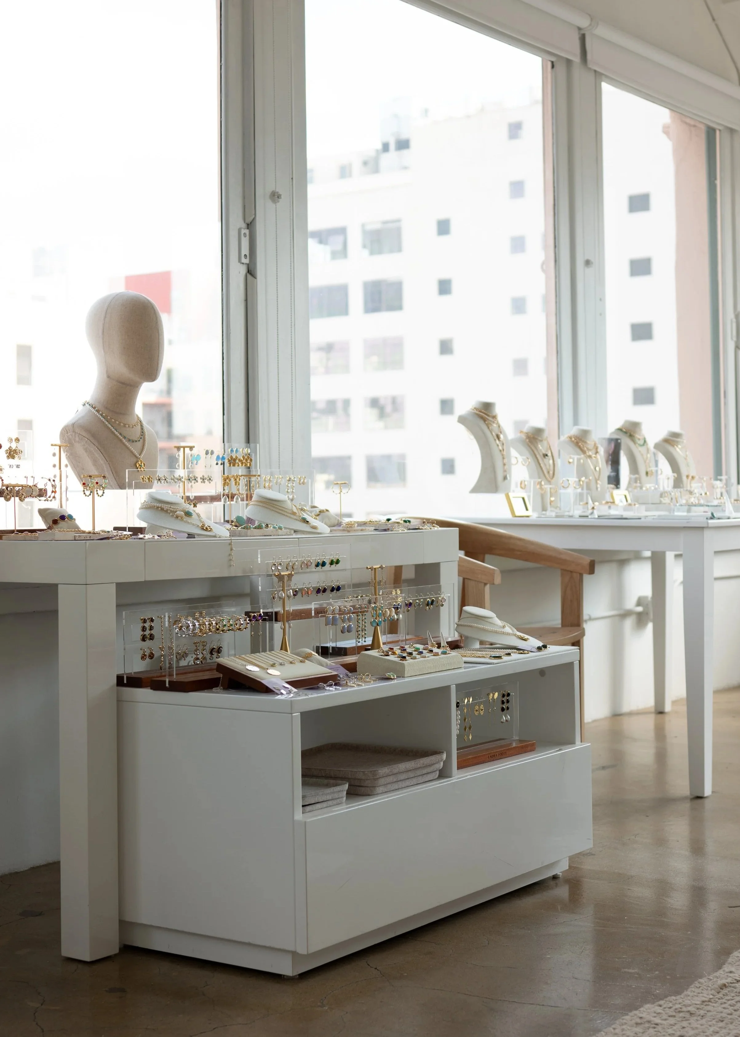 Display of jewelry including necklaces, earrings, and rings on white tables and stands inside a store with large windows.