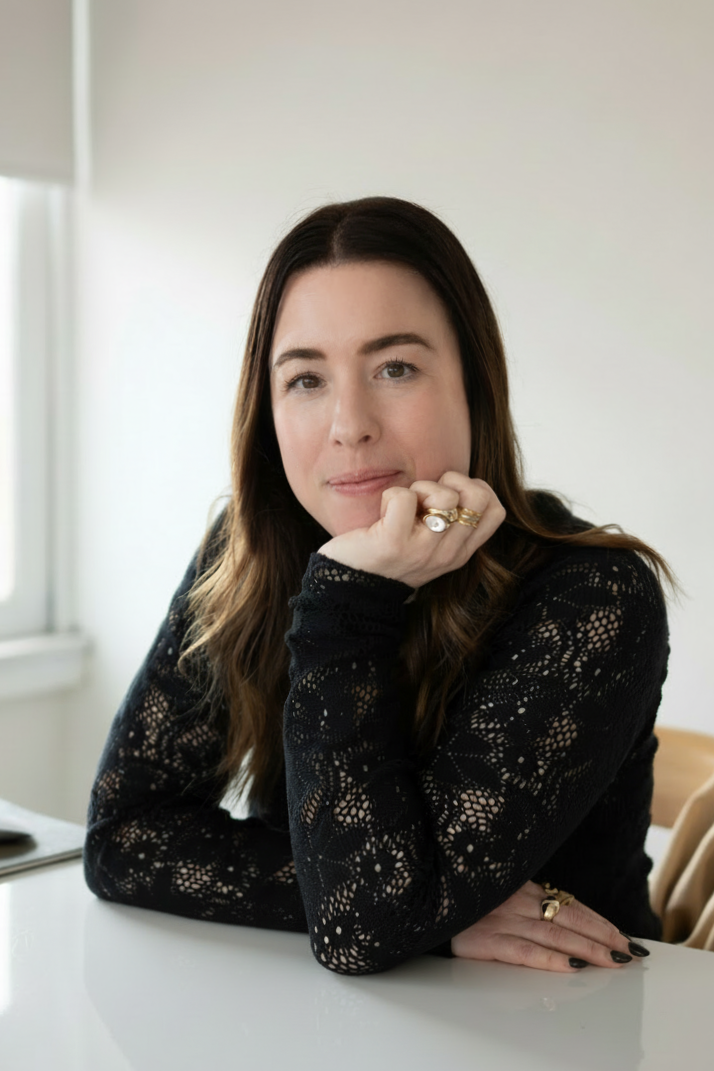 A woman with long brown hair sitting at a white table, resting her chin on her hand, wearing a black lace top, rings, and nail polish in a bright room with a window.