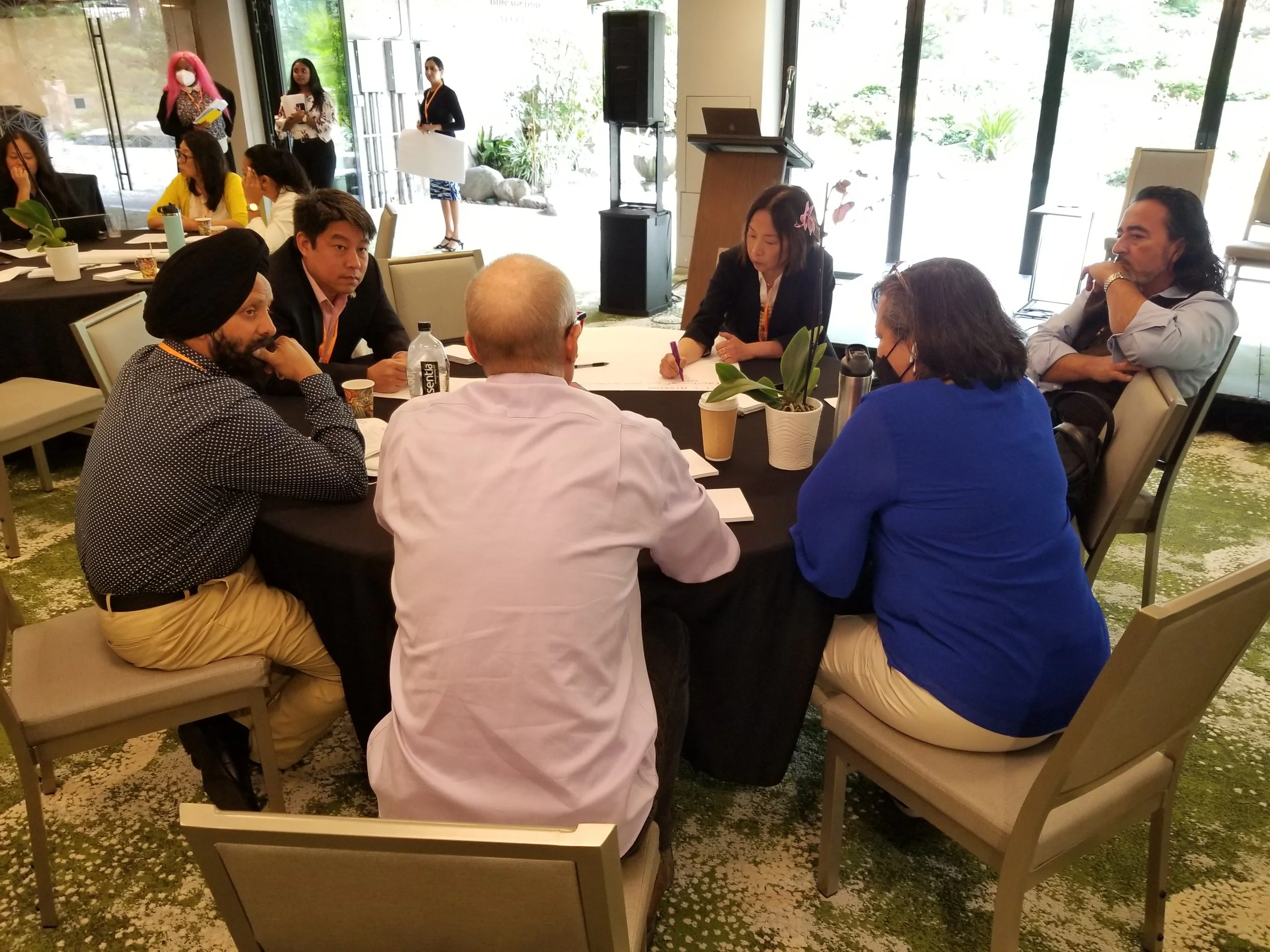 A small group of participants sits around a round table in a conference space, engaged in discussion and note-taking during an AAPI Against Gun Violence convening, with other attendees and a podium visible in the background.