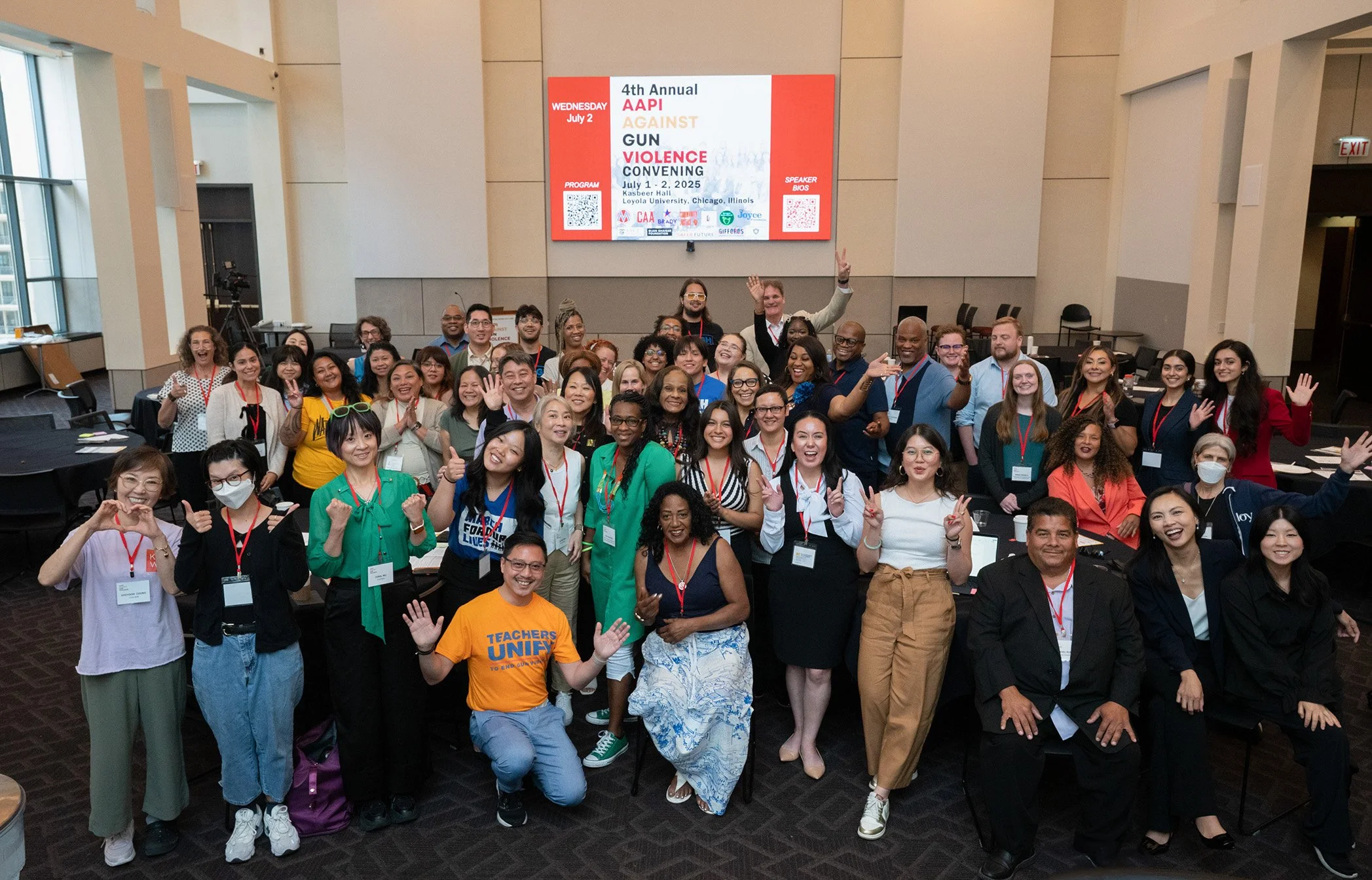 A large, diverse group of participants at an AAPI Against Gun Violence convening stands together in a conference room, smiling and waving, with an event screen visible behind them.