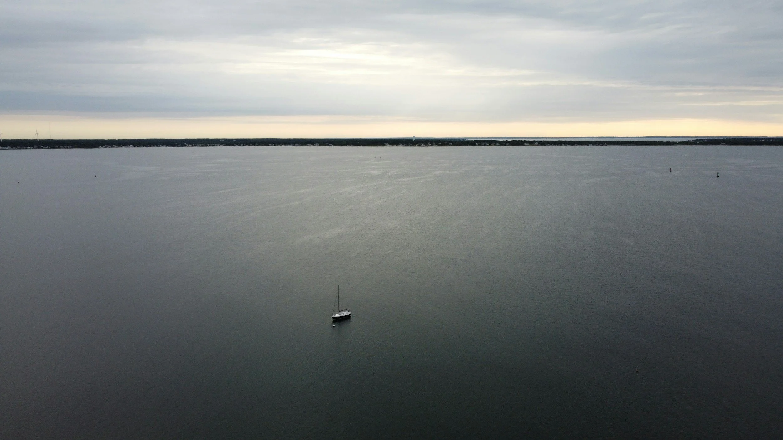 A solitary sailboat on a large, calm body of water under an overcast sky with a distant shoreline and a few small buoys or boats.