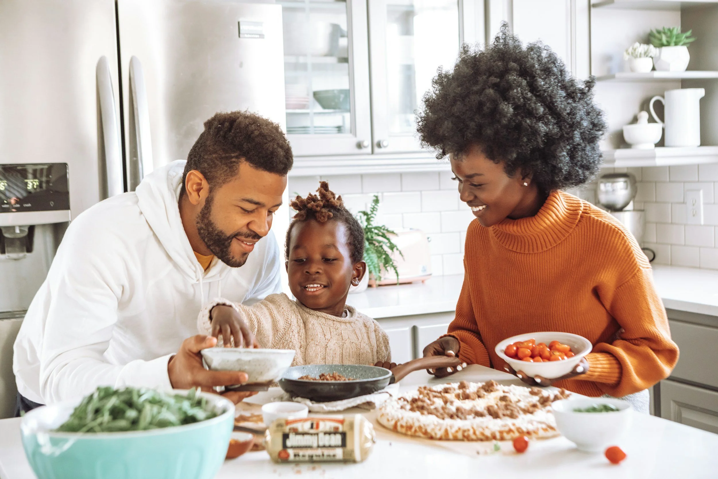 A family of three making and enjoying pizza together in a bright kitchen. The father, mother, and daughter are smiling and adding toppings to the pizza. The kitchen counter has bowls of cherry tomatoes, greens, and other ingredients.