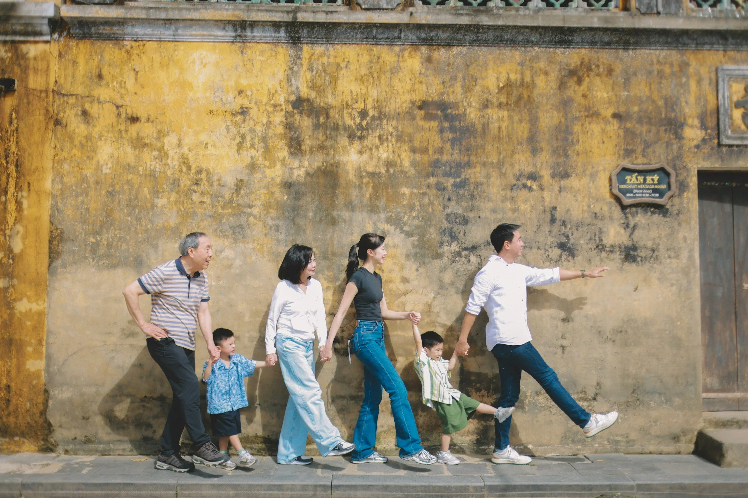 A multigenerational family walking hand-in-hand along a sidewalk in front of an old yellow textured wall.