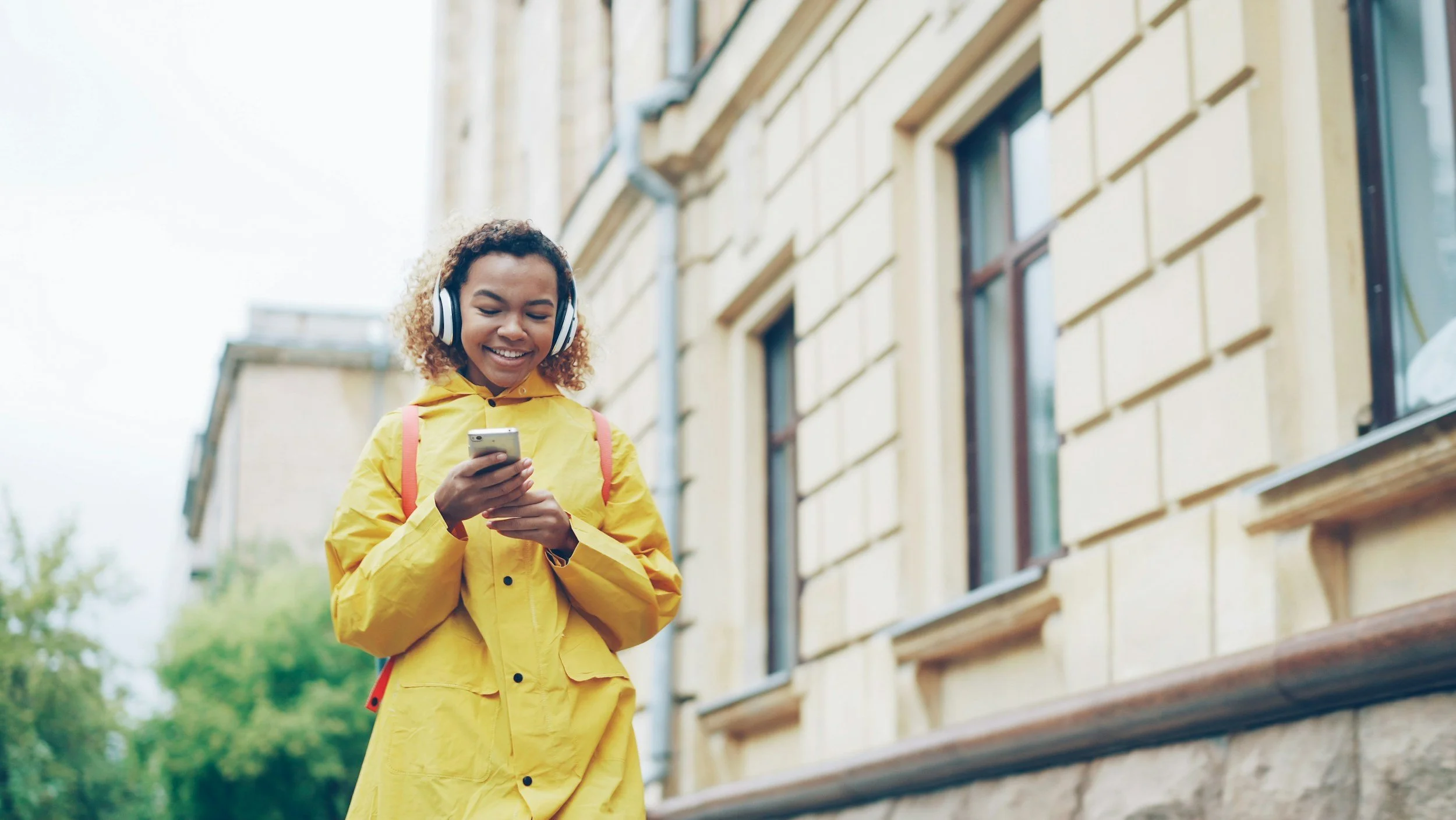 Young woman in yellow raincoat with backpack, listening to music with headphones, using smartphone outdoors in urban setting.