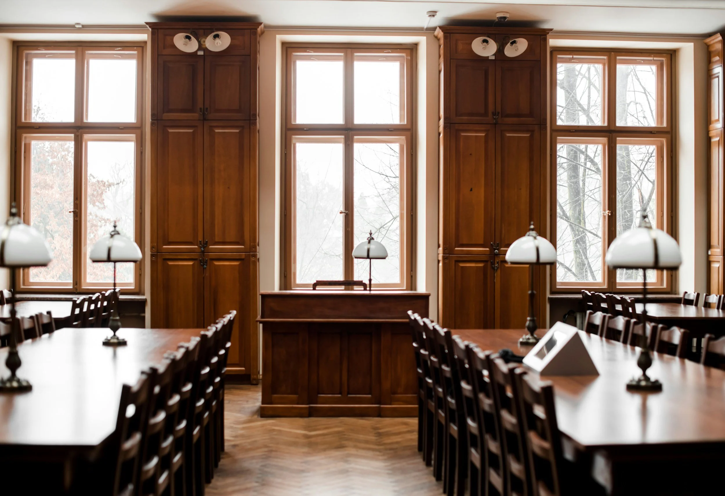 A dining room with large wooden tables, chairs, and lamps, featuring tall windows with wood framing and view of trees outside.