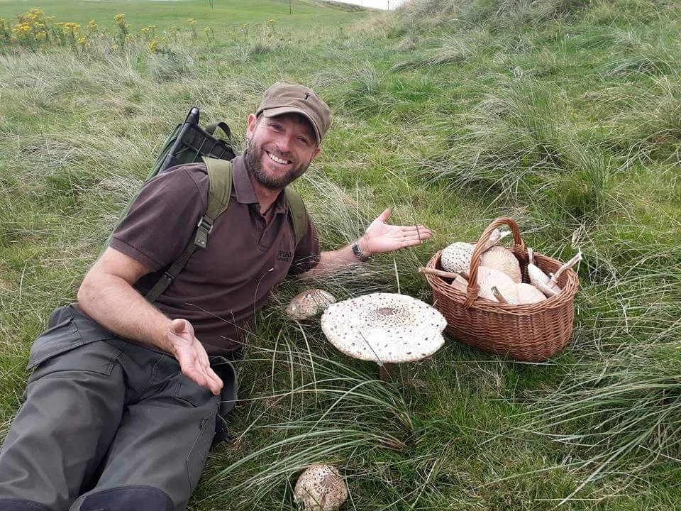 Man smiling with a backpack near a basket of mushrooms and some mushrooms on the grass in a grassy outdoor area.