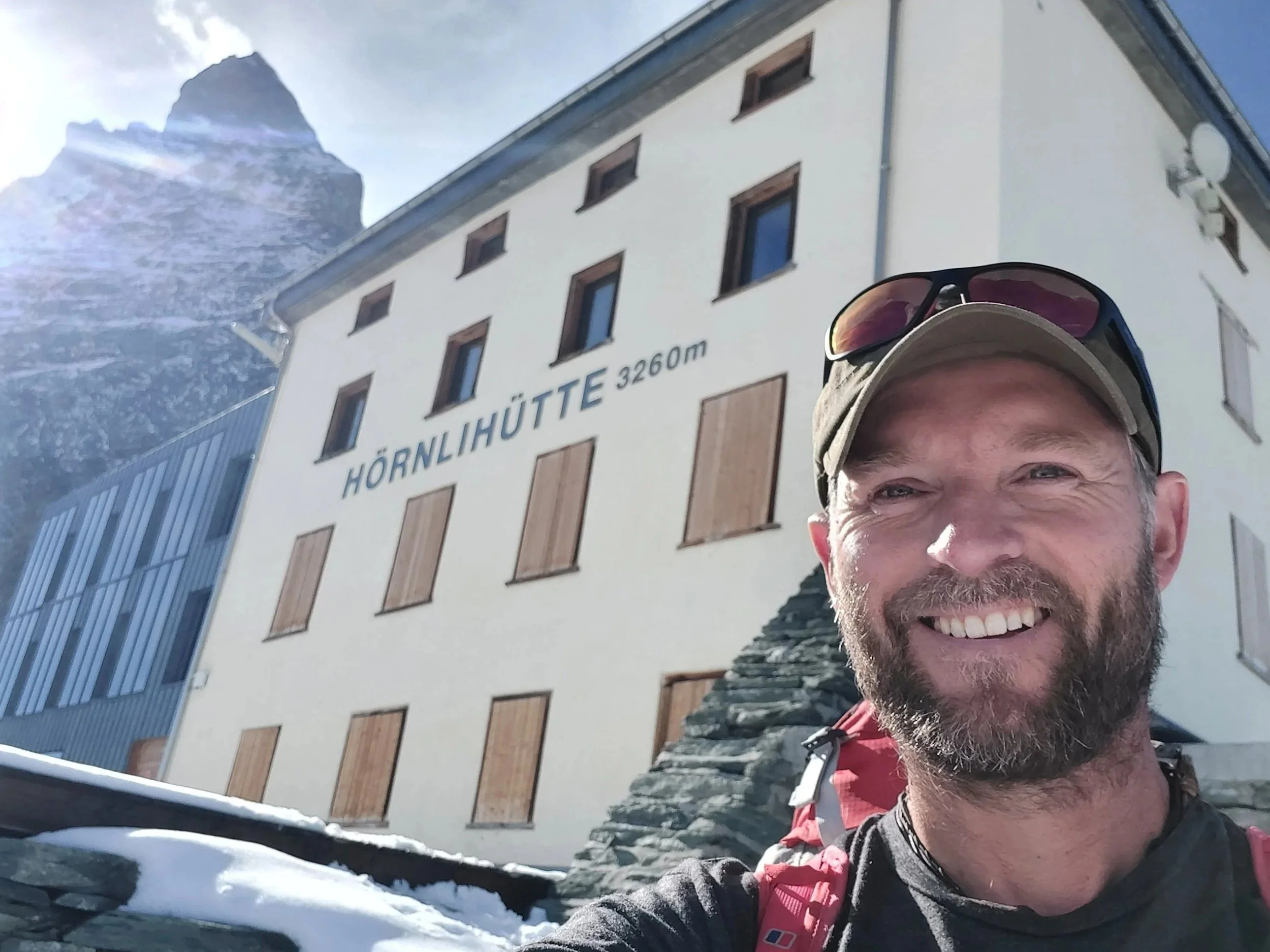 Smiling man in outdoor gear taking a selfie in front of Hörnlihütte at 3,260 meters in the mountains with a snowy landscape and mountain peak in the background.