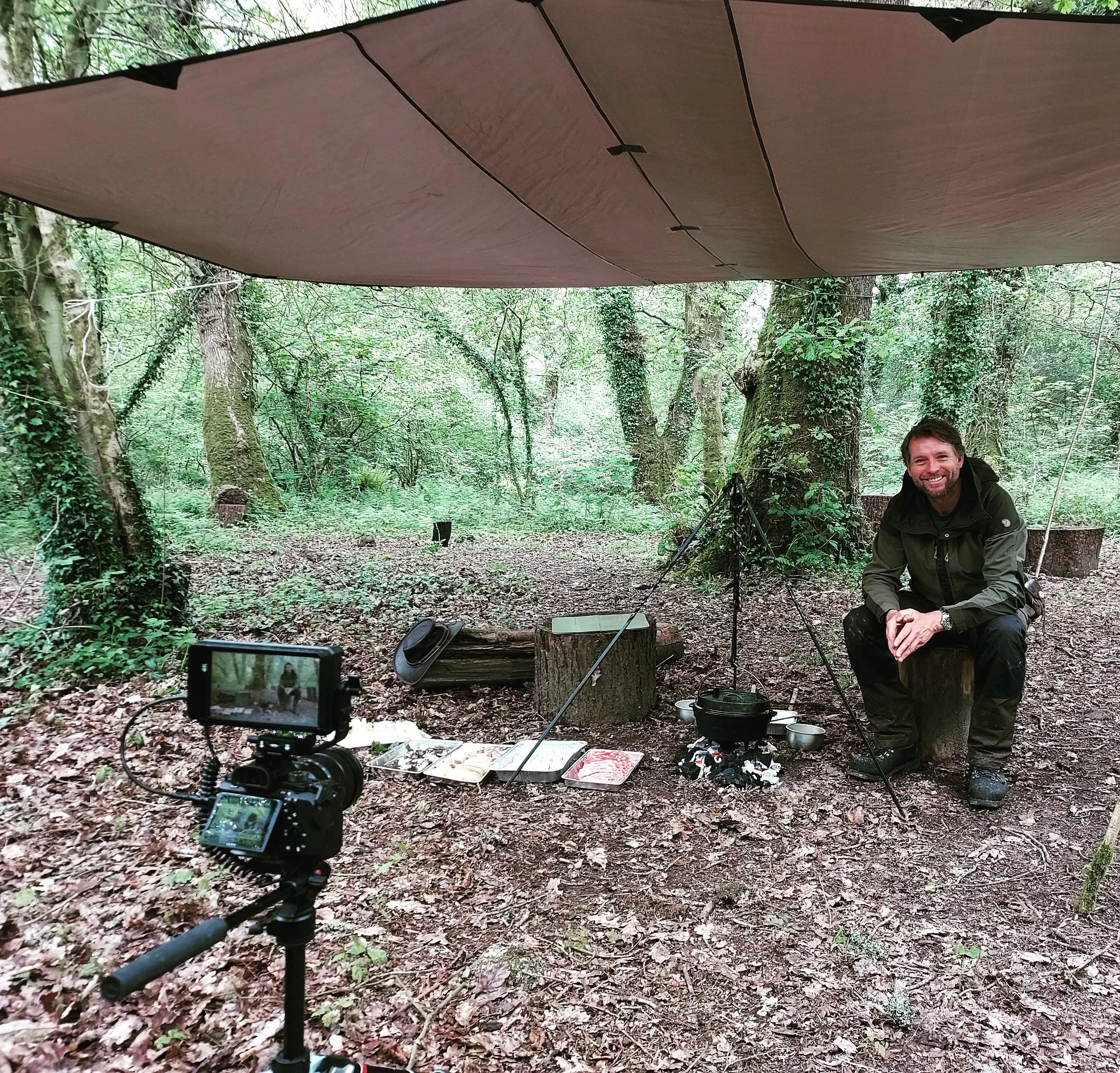 A man sits on a tree stump in a forest, smiling, with a filming setup that includes a camera on a tripod, a tarp overhead, and camping equipment like a hanging pot over a small campfire, with food in plastic containers on the ground.