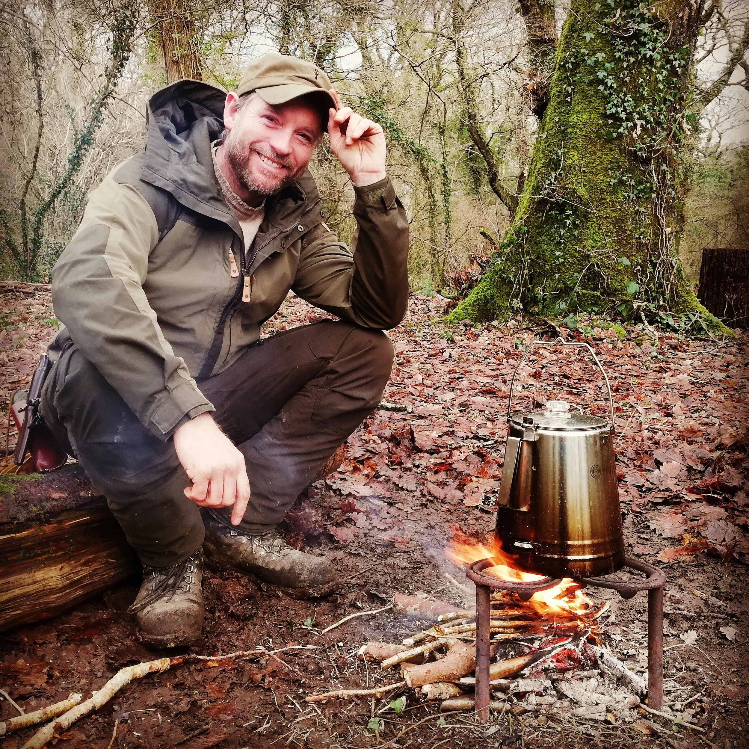 A man dressed in outdoor gear squats next to a campfire in a wooded area, with a metal kettle hanging over the fire.