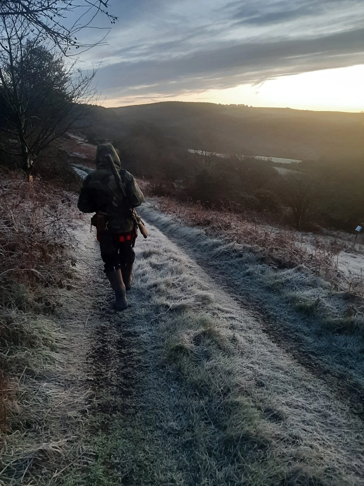 A person walking along a frost-covered dirt trail in a rural, hilly landscape during sunrise or sunset, carrying a rifle slung over their shoulder and dressed in camouflage clothing.