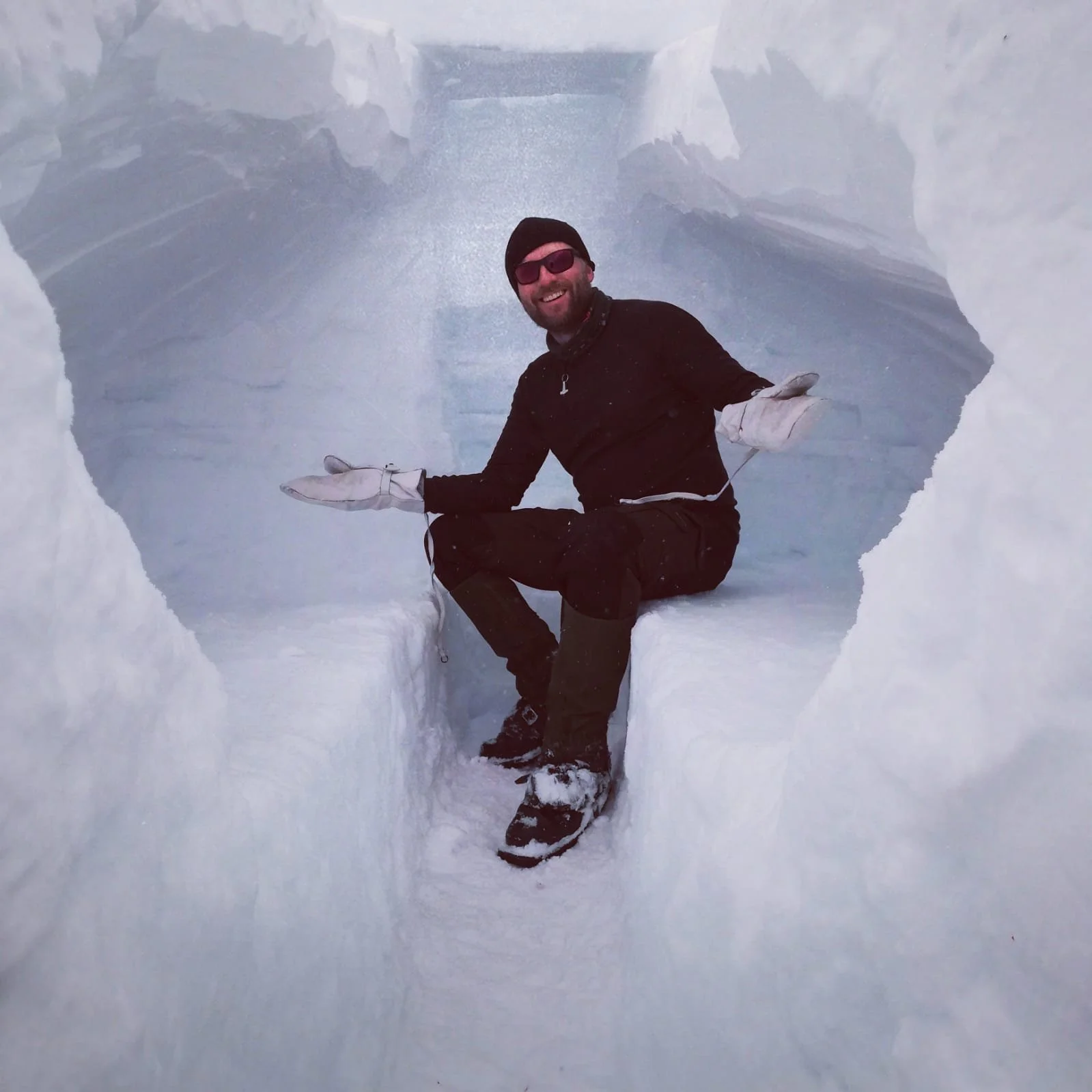 A man sitting inside an ice cave with walls of blue and white ice, smiling and wearing winter clothes and sunglasses.