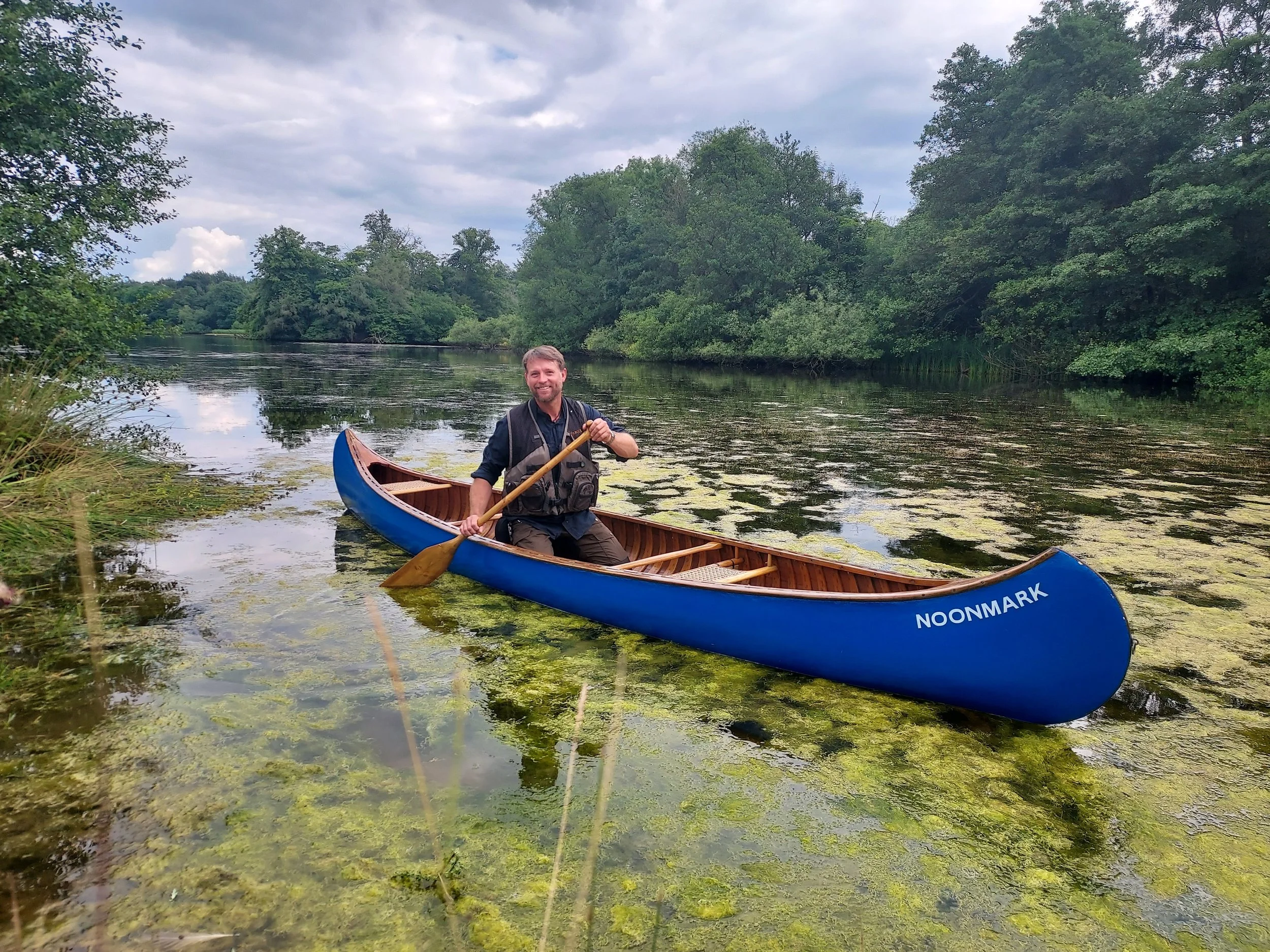 A man is sitting in a blue canoe on a river, holding a paddle, surrounded by greenery and trees, with a cloudy sky overhead.