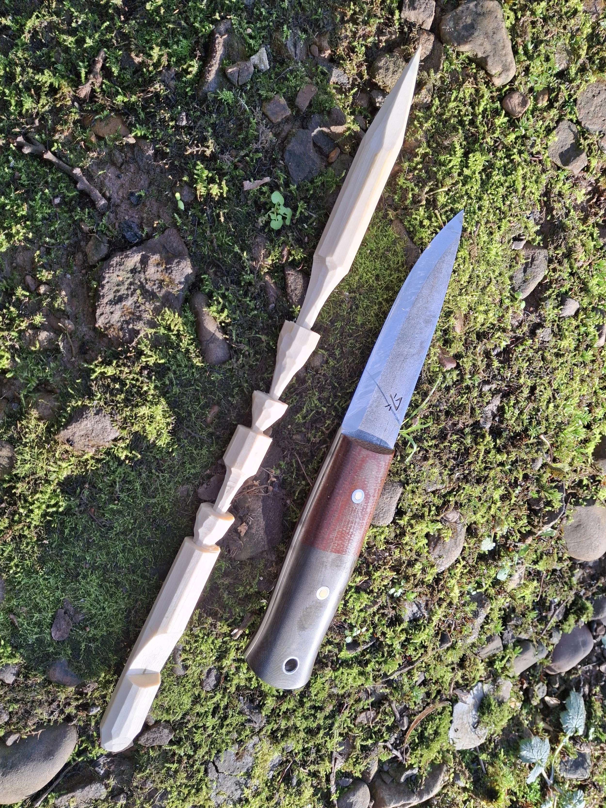 A crafting knife and a wooden blade on a grassy ground with small rocks and moss.