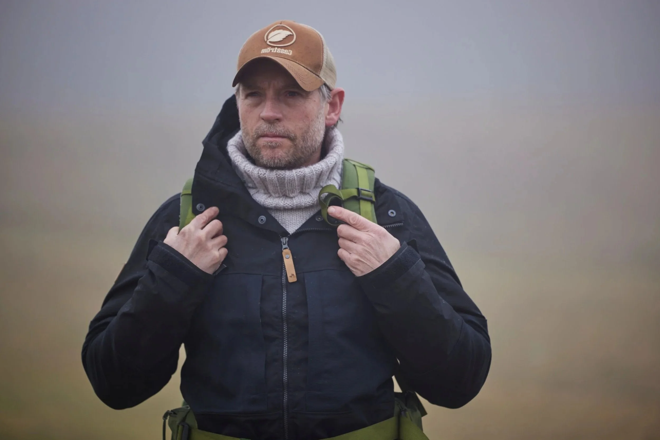 A man with a beard dressed in outdoor clothing, including a black jacket, gray turtleneck, and brown cap, carrying a green backpack in a foggy outdoor setting.