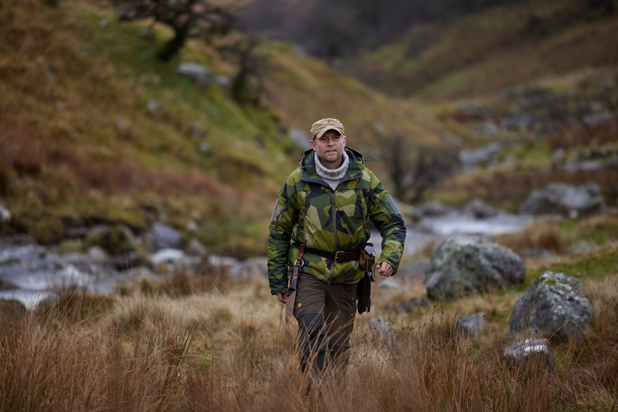 A man hiking through a rugged, natural landscape with grassy and rocky terrain, and a stream running through the background.