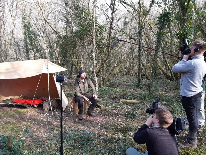 Film crew filming an interview with a man sitting on a tree stump in a wooded outdoor setting, with filming equipment and a tent nearby.