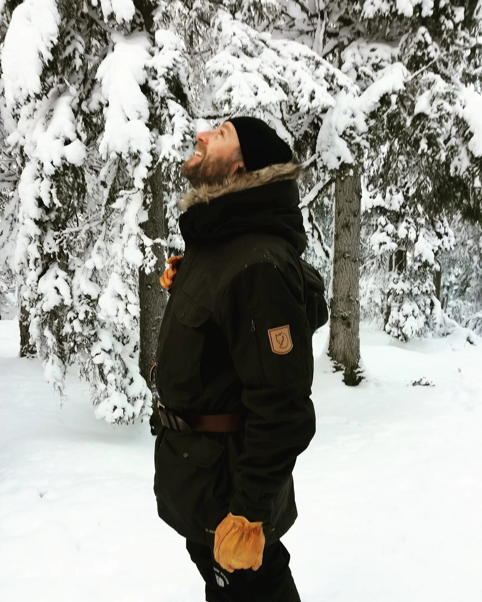 A man standing outdoors in a snow-covered forest, smiling and looking upwards, wearing a black jacket, black beanie, and yellow gloves.