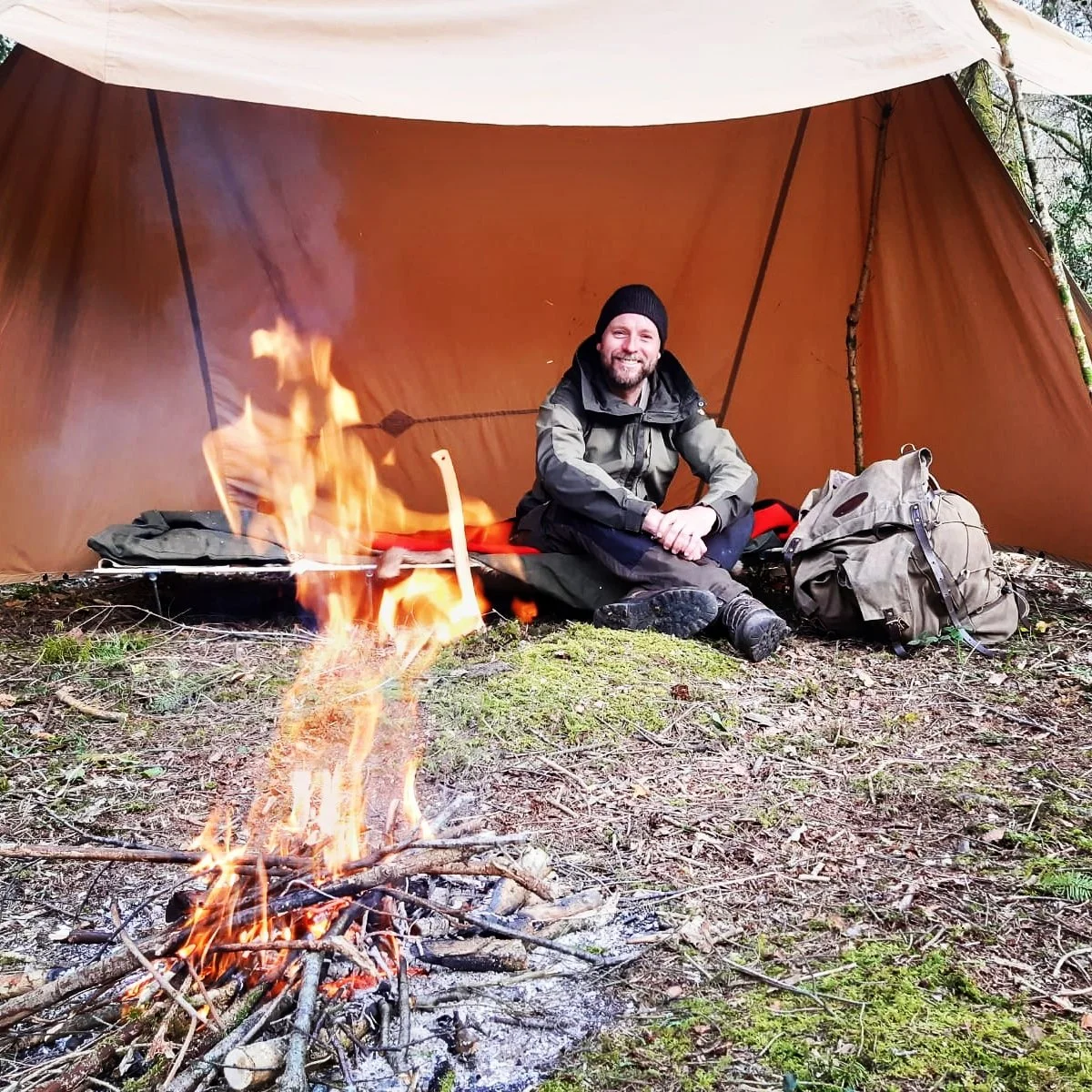 A man sitting inside a camping tent with a campfire burning in front of him in a wooded area. He is smiling, wearing outdoor gear, and there are backpacks nearby.