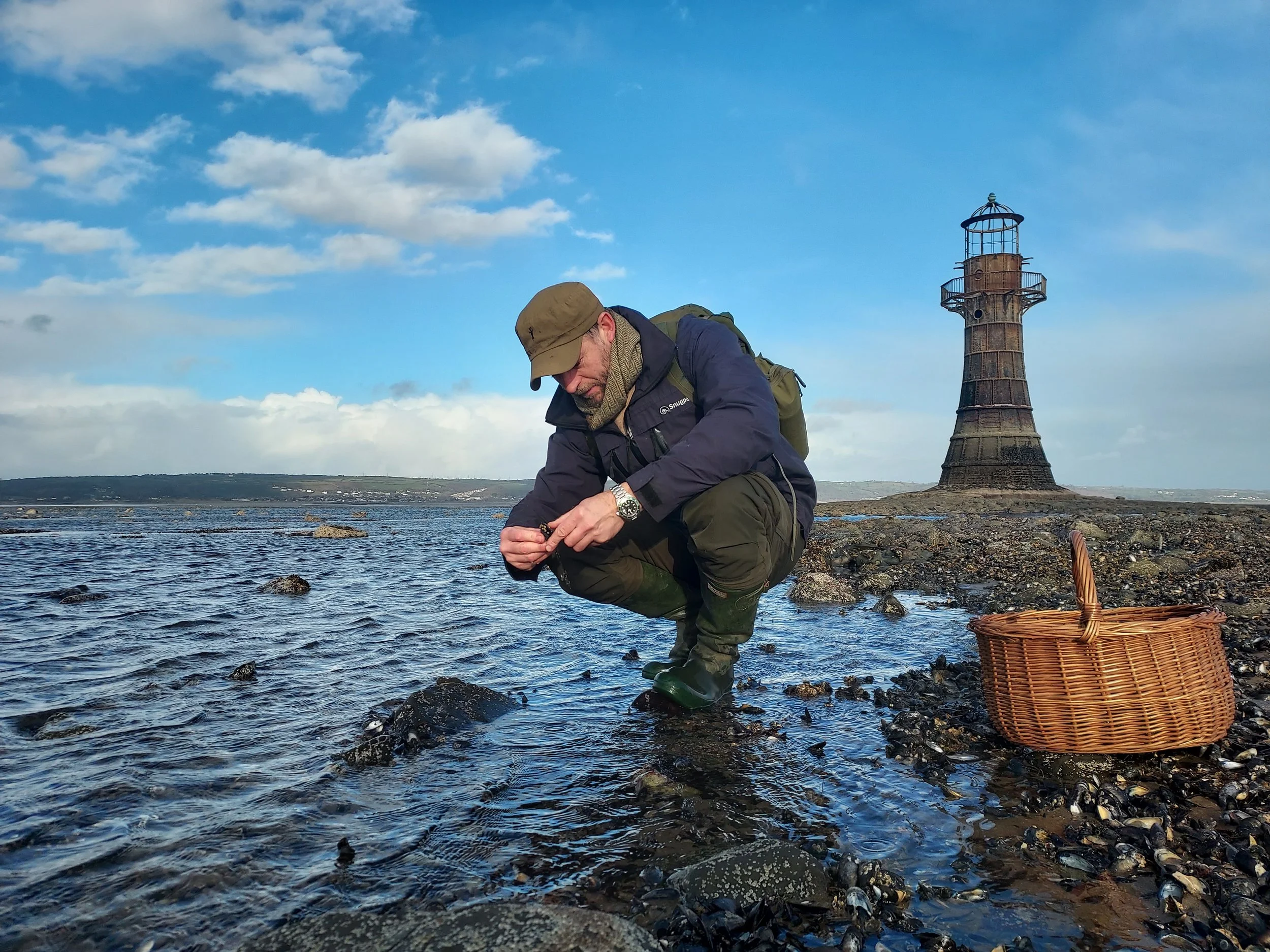 Man wearing outdoor gear and rubber boots crouching on rocky shoreline, holding something in his hands with a basket nearby; lighthouse in the background and partly cloudy sky.