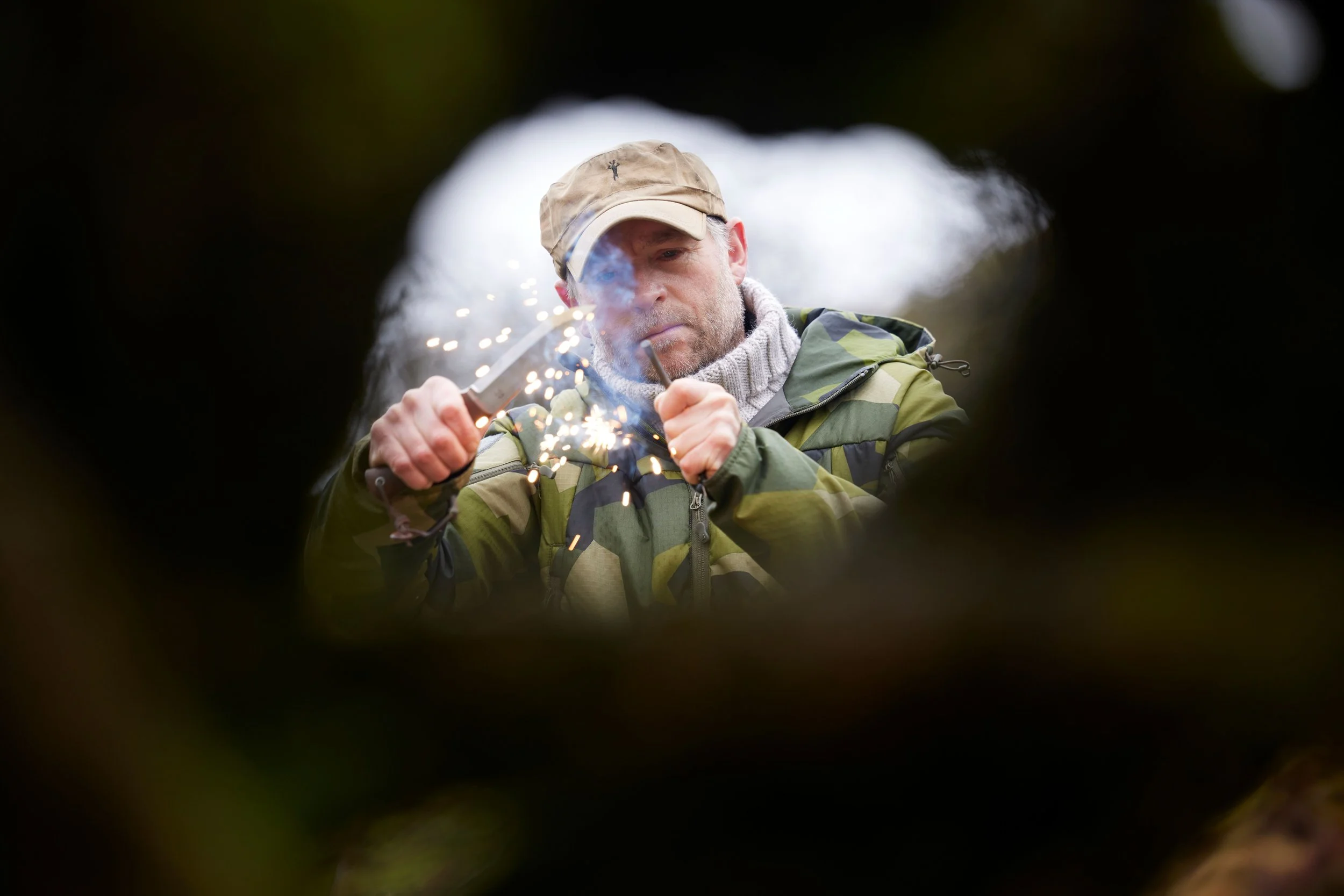 A man wearing camouflage jacket and beige cap, seen through a hole, is lighting a fuse with a sparkler, with sparks and smoke around him.