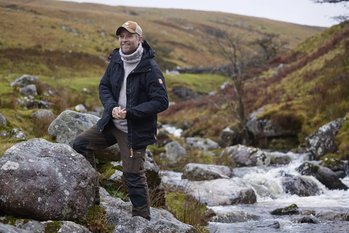 A man wearing outdoor clothing, including a baseball cap, gray turtleneck, and black jacket, stands on rocks by a small stream in a hilly landscape with sparse trees and yellow-brown vegetation.