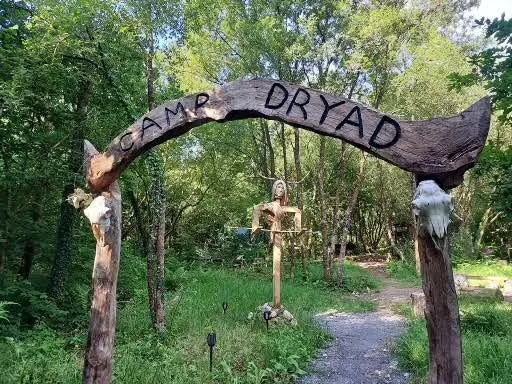 A wooden archway with the words  "CAMP DRYAD" written on it, situated in a forested area with green trees and a dirt trail.