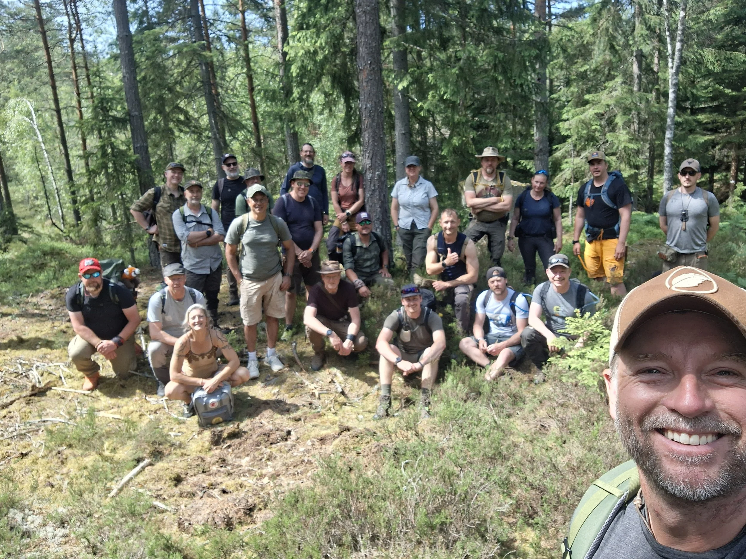 A group of hikers in a forest taking a group photo, with some standing and some kneeling, surrounded by tall trees and green foliage.