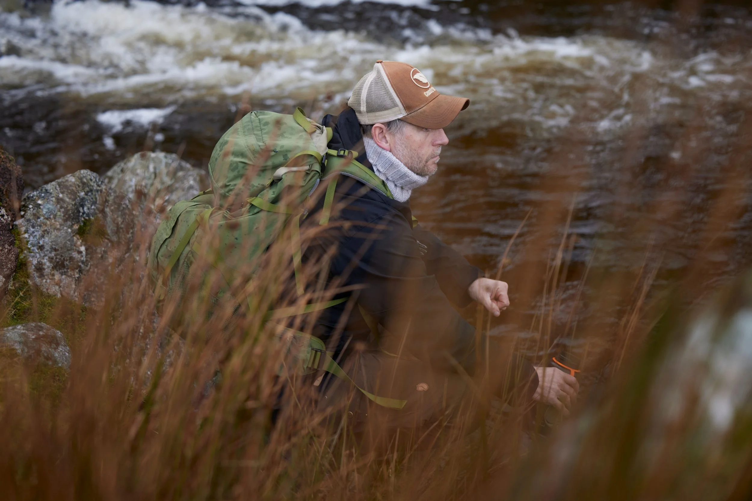 A man with a backpack sitting by a creek, holding a cup, surrounded by tall grass.