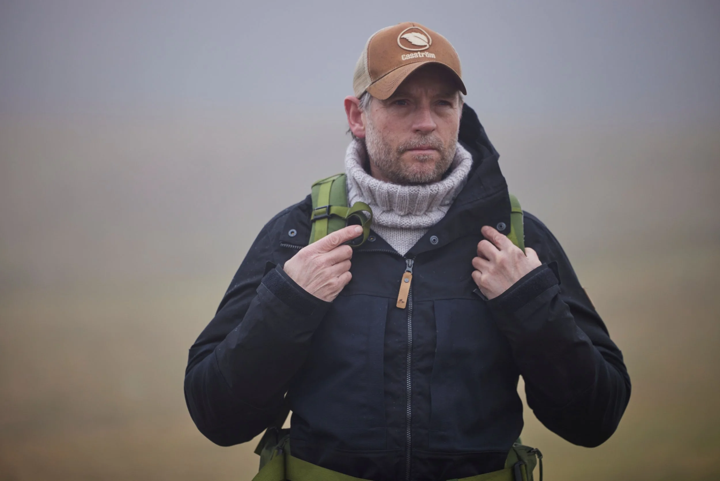 A man with a beard wearing a brown cap, black jacket, gray turtleneck, and backpack, standing outdoors in foggy weather.