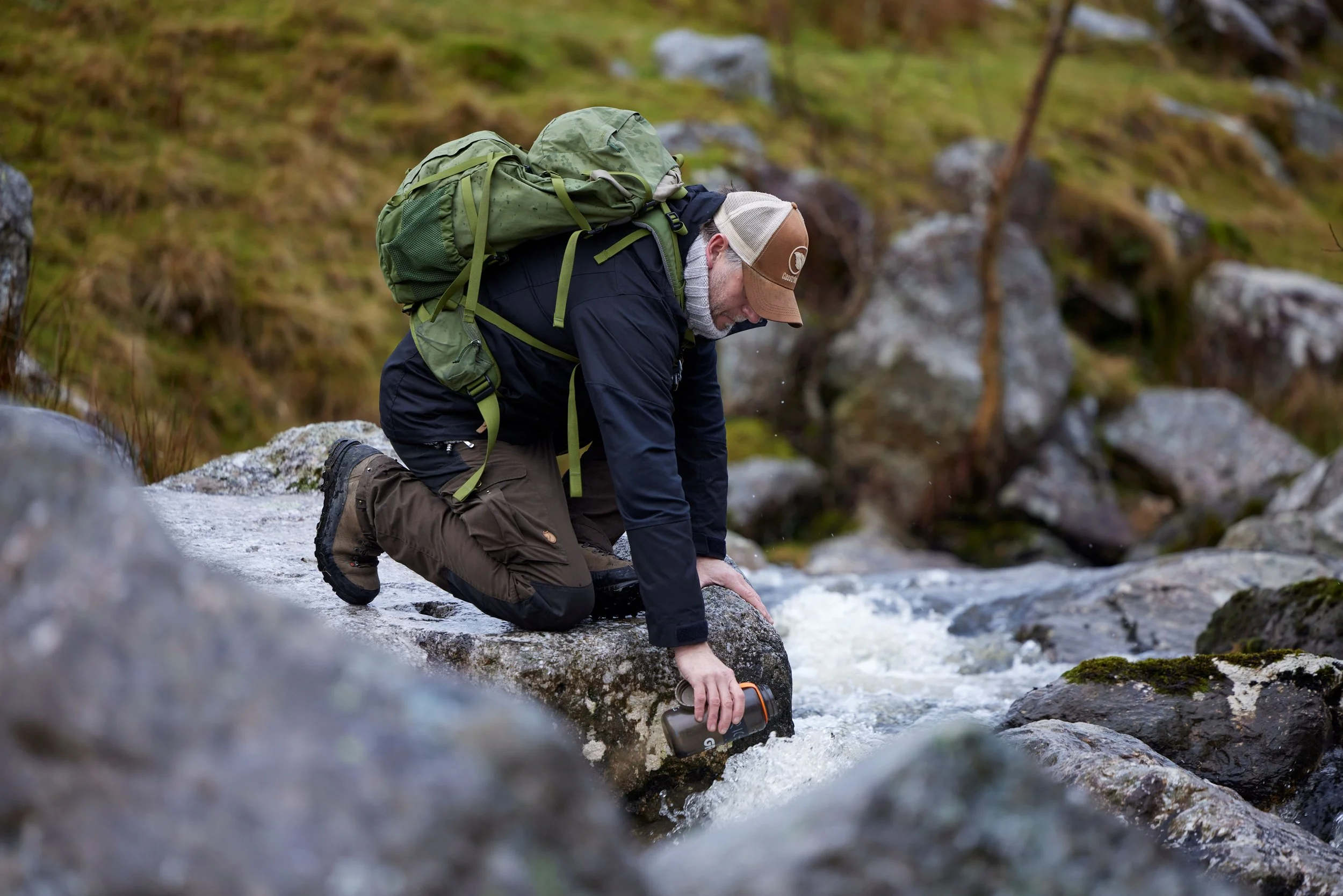A man dressed in outdoor clothing and backpack is kneeling by a stream and pouring water into a container.