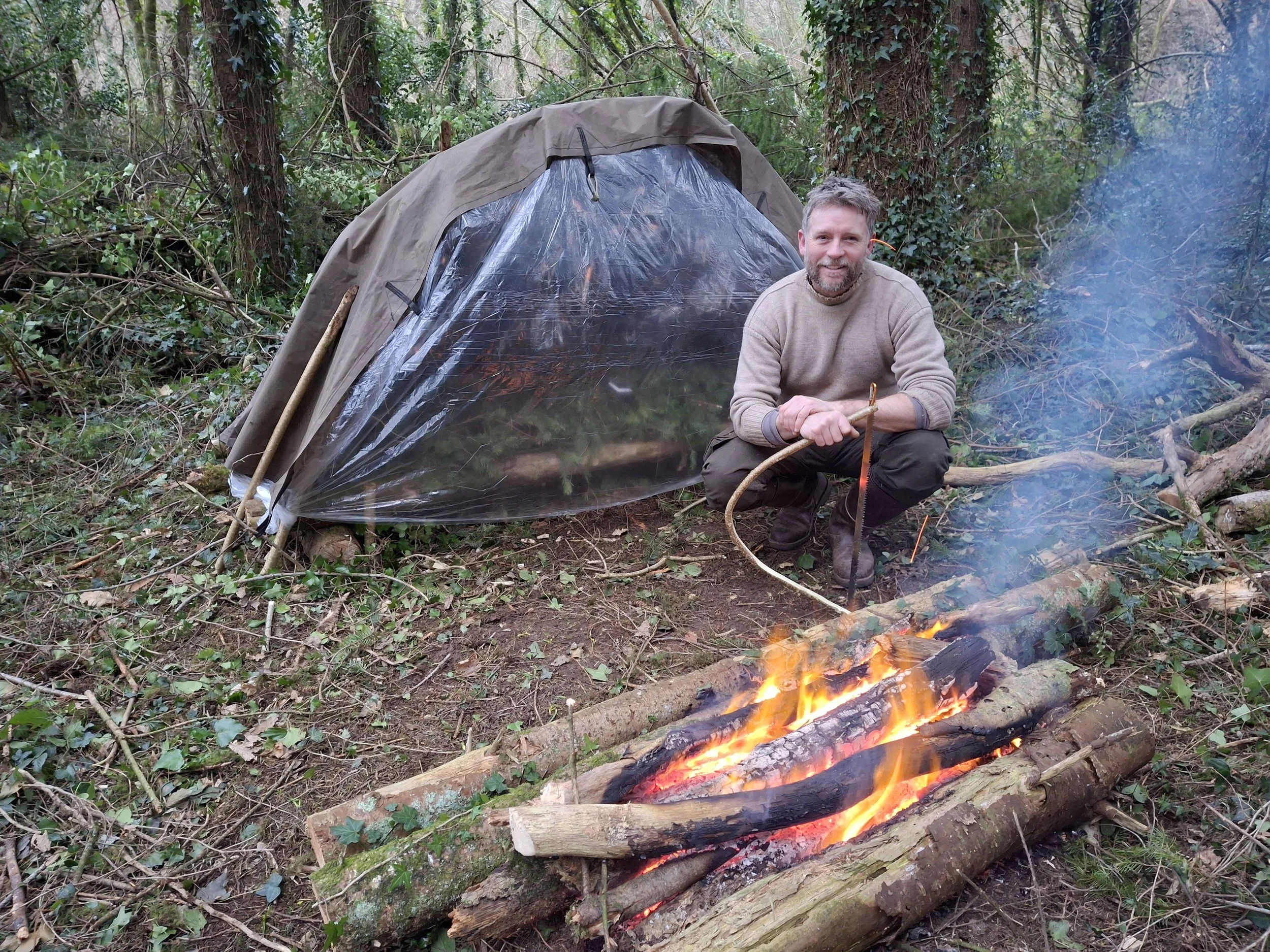 Man crouching next to a campfire with a tent in the background in a wooded area.