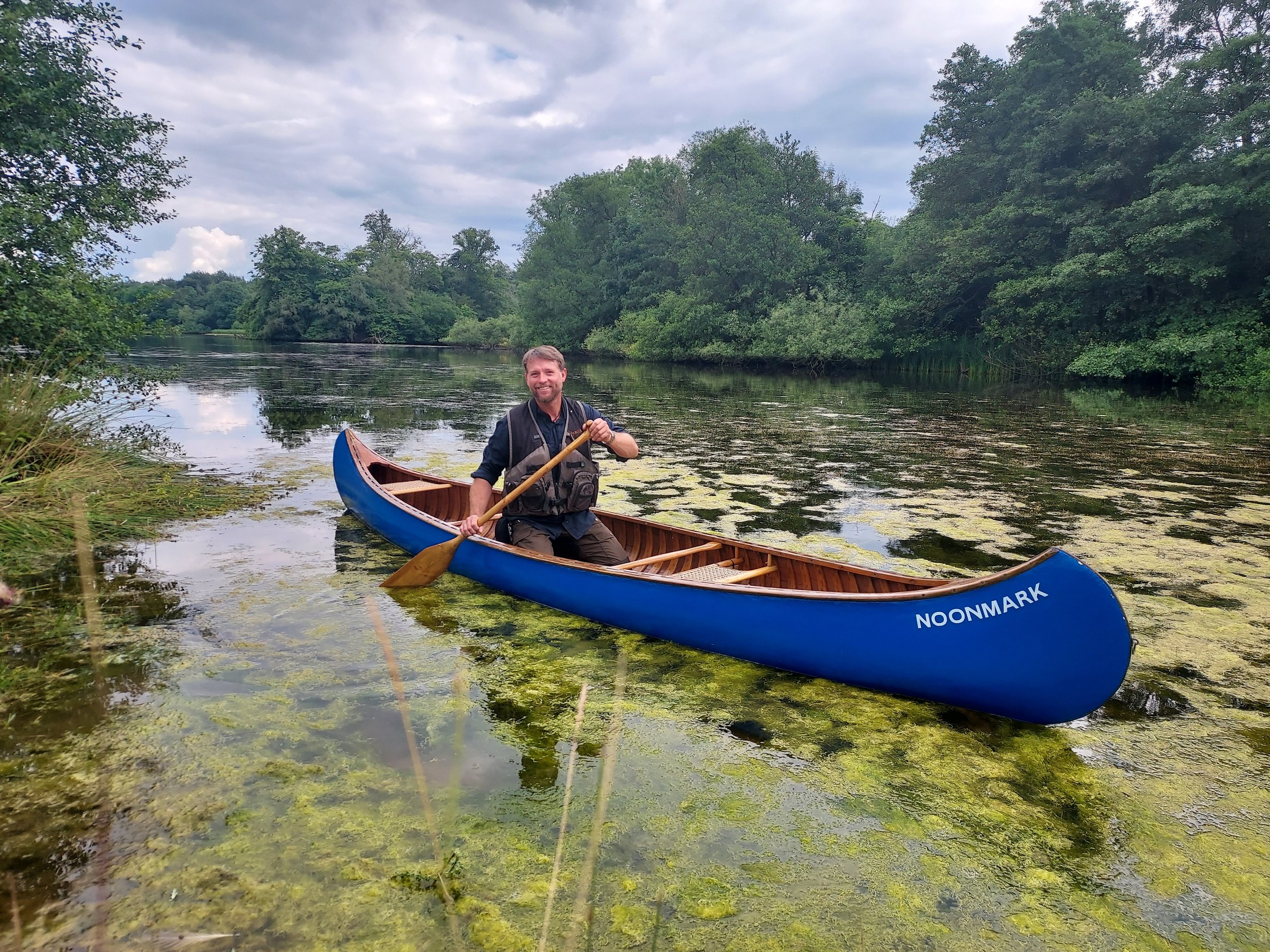 A man smiling and sitting in a blue canoe on a calm, algae-covered river surrounded by green trees under a cloudy sky.