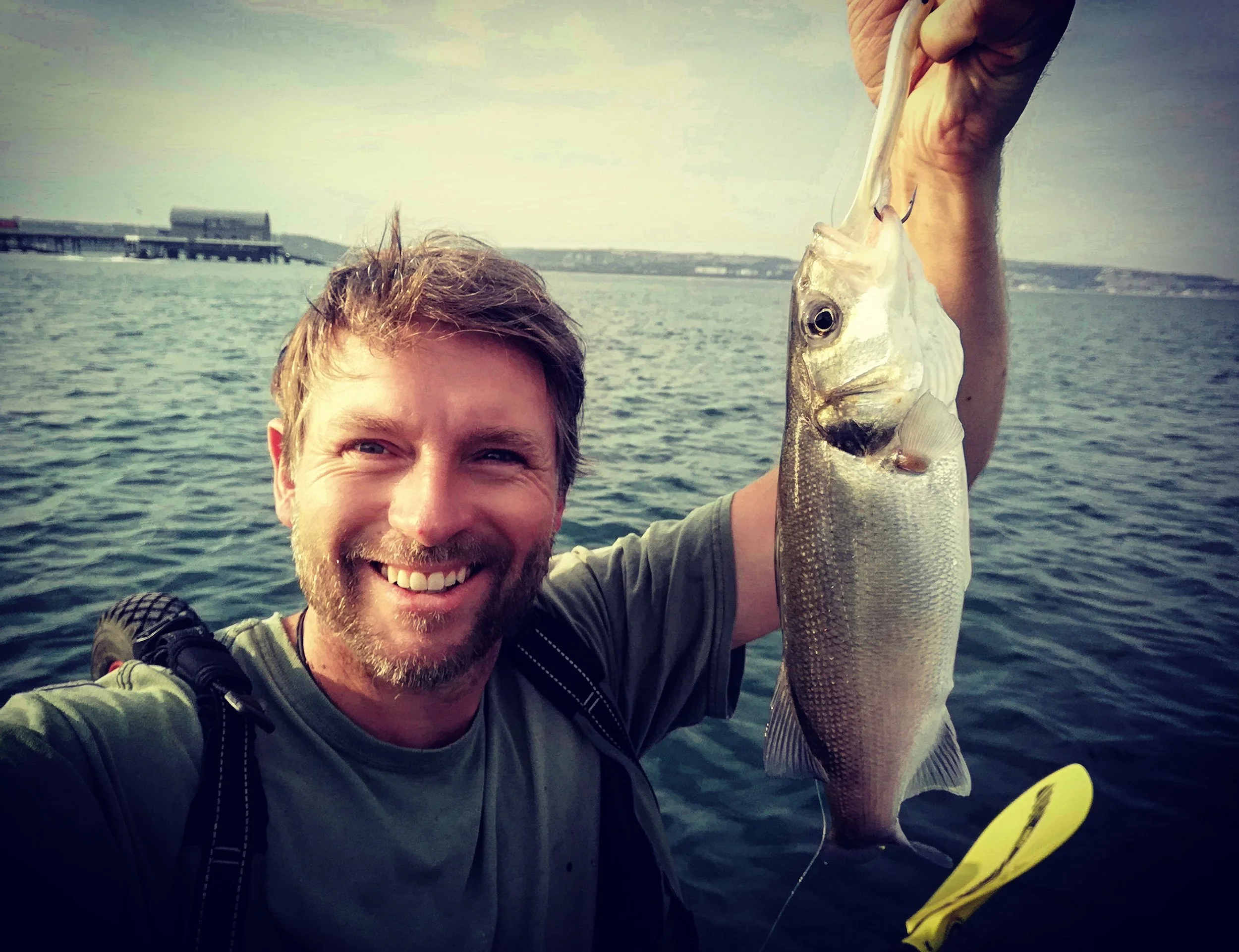 A smiling man holding a fish he caught over a body of water, with a distant shoreline and structures in the background.
