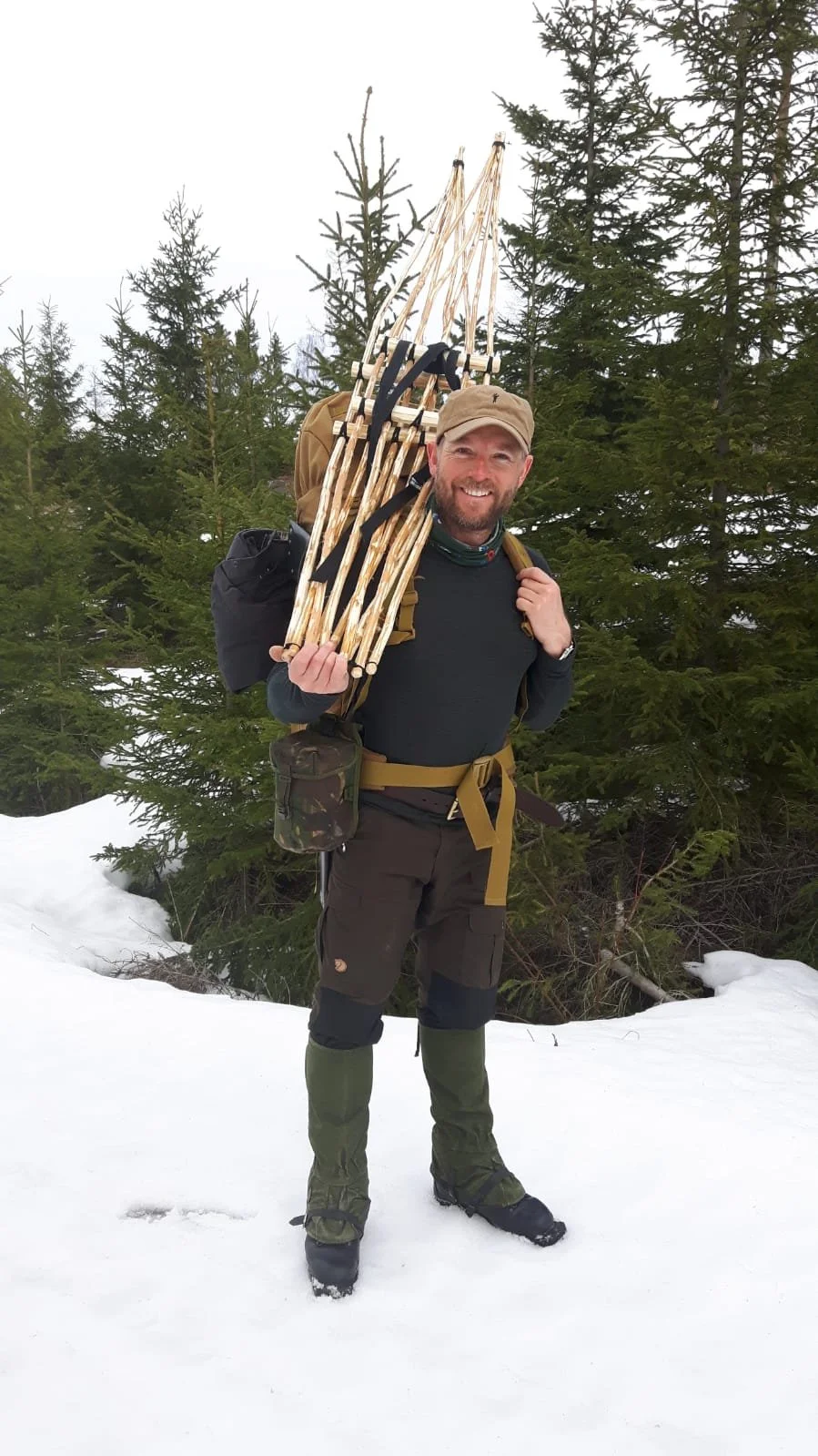 A man with a beard smiling, carrying a large bundle of walking sticks on his shoulder, standing in snow with evergreen trees in the background.